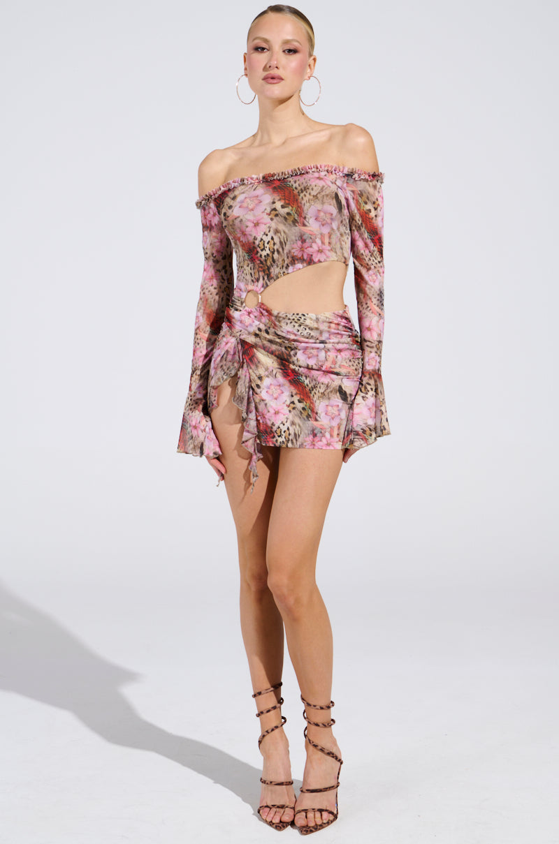 Wearing the MIAMI NIGHTS MINI DRESS with its bodycon silhouette and floral off-the-shoulder design, complete with a cutout tie detail and long sleeves, they pose confidently. Hoop earrings and strappy high heels enhance the look against a plain background.