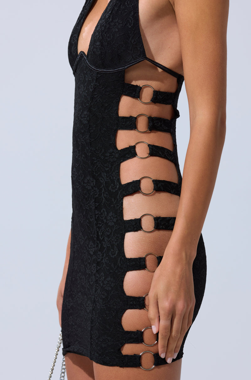 Wearing the NOT FOR EVERYONE CUTOUT LACE MINI DRESS, a black floral lace bodycon dress with cutout sides and metal ring details, someone stands against a light background, highlighting the bold silhouette.