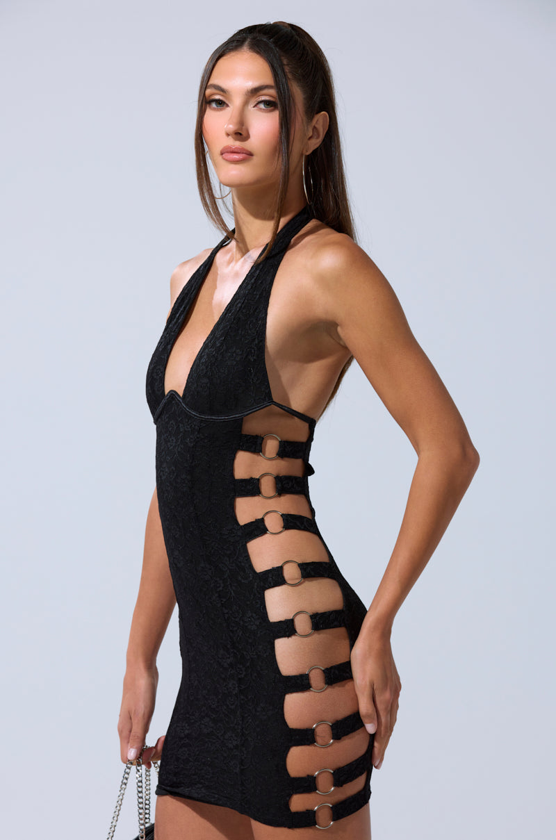 A woman wears the NOT FOR EVERYONE CUTOUT LACE MINI DRESS, a black halter mini with side cut-outs and ring details, standing against a plain light background and holding a chain-strap accessory by her side.