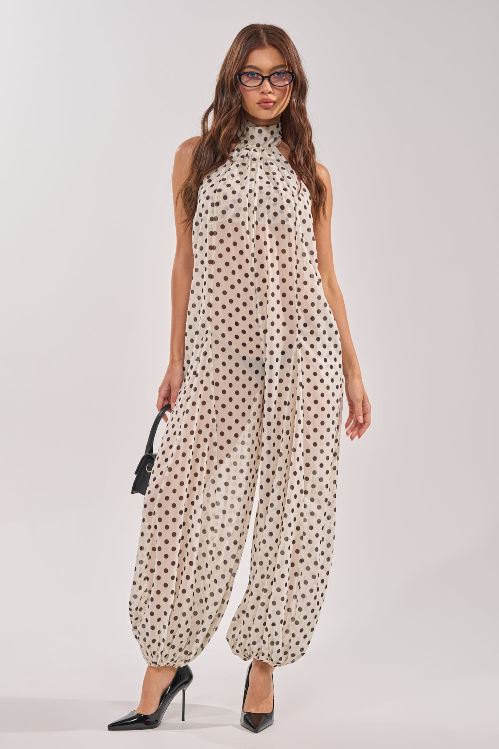A woman in glasses models the GIRLY POP POLKA DOT JUMPSUIT—beige with white polka dots and gathered pant legs. She accessorizes with a small black handbag and black pointed heels, set against a plain backdrop. 0
