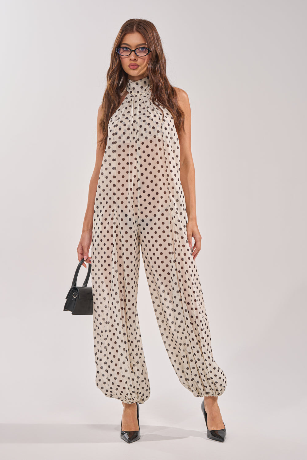 A woman stylishly poses against a white backdrop in the GIRLY POP POLKA DOT JUMPSUIT, paired with black heels, glasses, and a small black handbag. 1