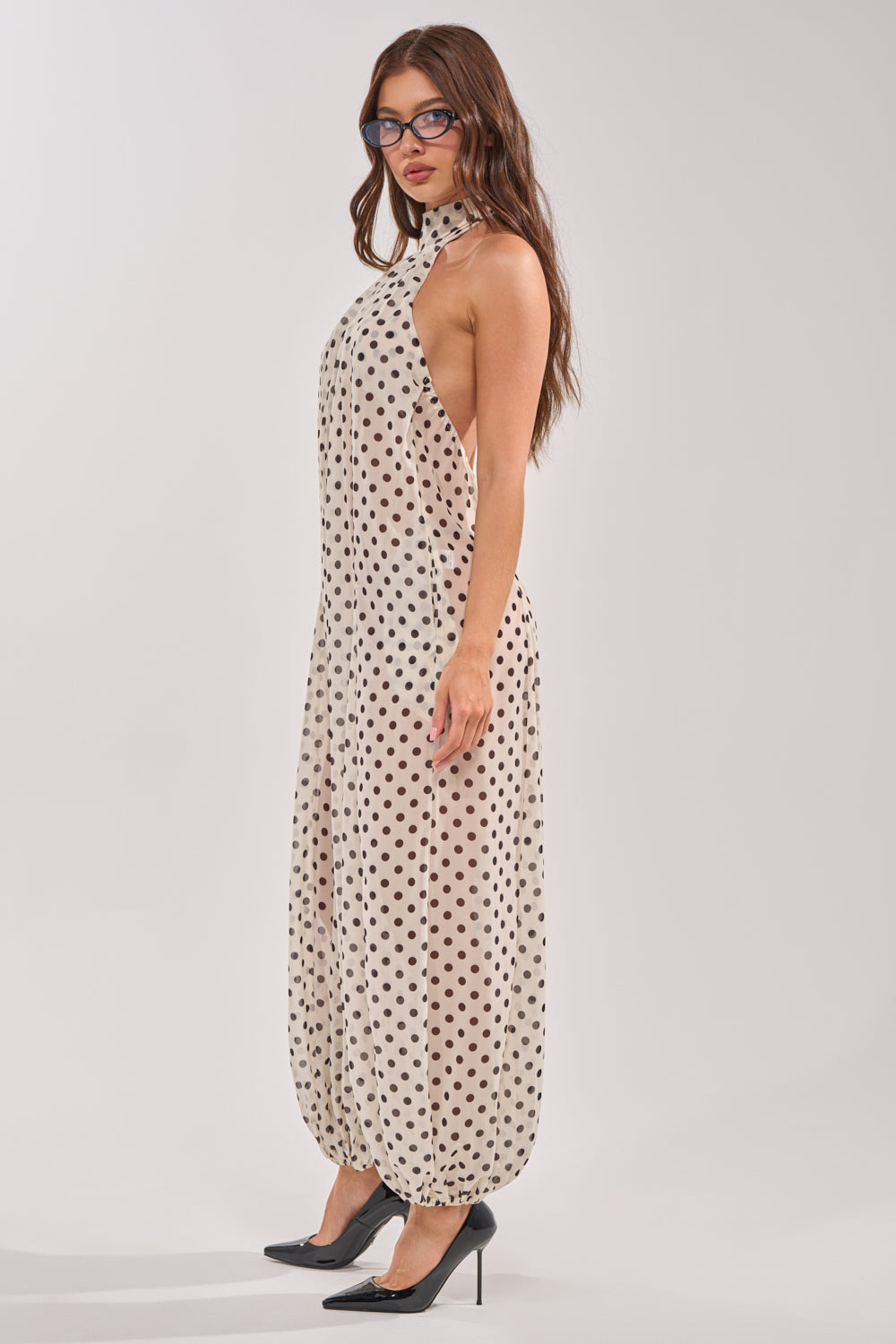 A woman in glasses models the GIRLY POP POLKA DOT JUMPSUIT, a chic cream sleeveless halter-neck jumpsuit with black polka dots and ankle length. She stands in profile against a plain background, wearing black high heels. 2