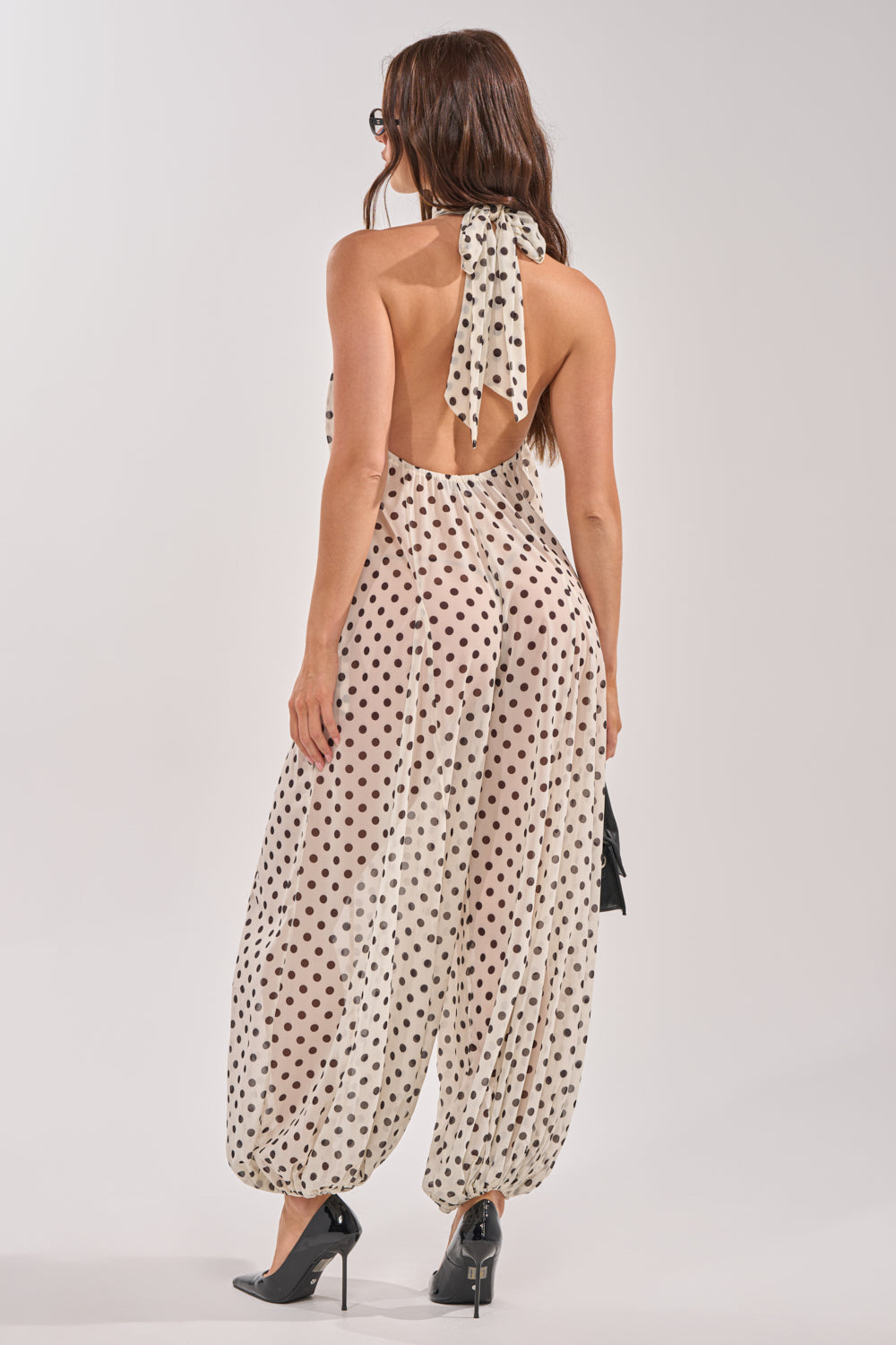 A woman with long brown hair and black heels models the GIRLY POP POLKA DOT JUMPSUIT—a light, backless halter-neck style with black dots—posing from behind to show its loose fit and neck bow detail. 3
