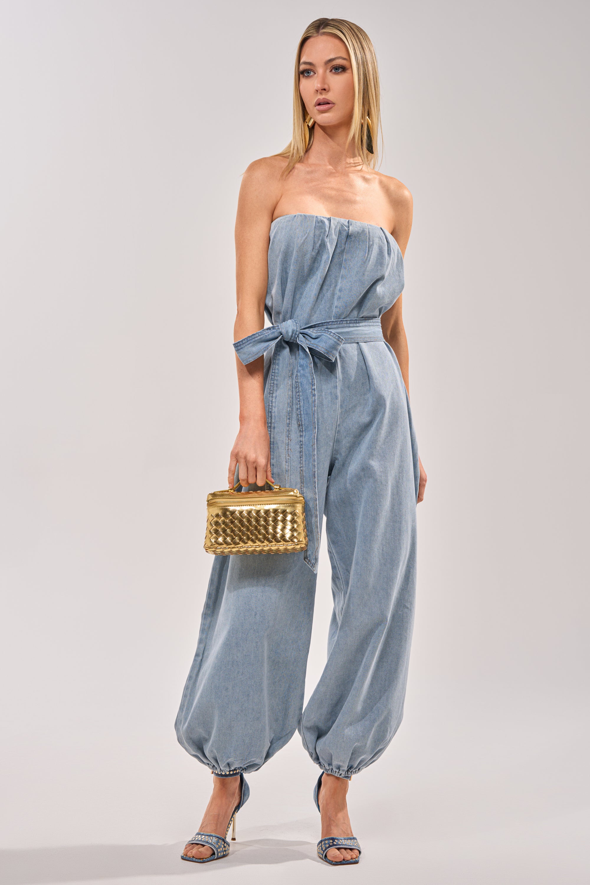 A woman stands against a plain background wearing the BREEZY DAY DENIM JUMPSUIT—a strapless, light-wash denim jumpsuit with gathered ankles—styled with blue heeled sandals and a gold textured handbag for a chic summer look. 1
