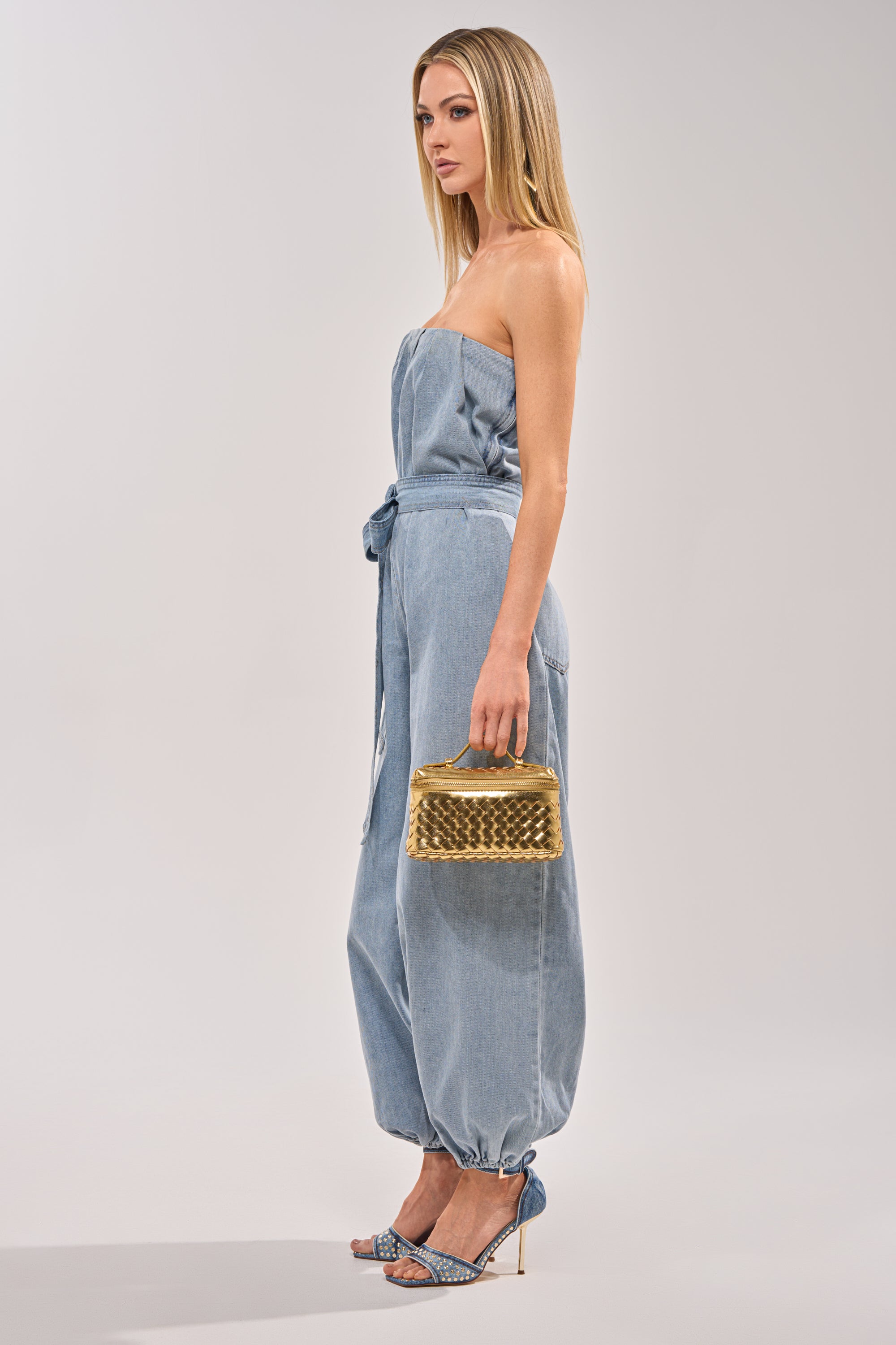 A woman stands in profile against a light gray background wearing the BREEZY DAY DENIM JUMPSUIT, a strapless light-wash style with a tied waist and gathered ankles. She accessorizes with blue heels and a small gold handbag. 2