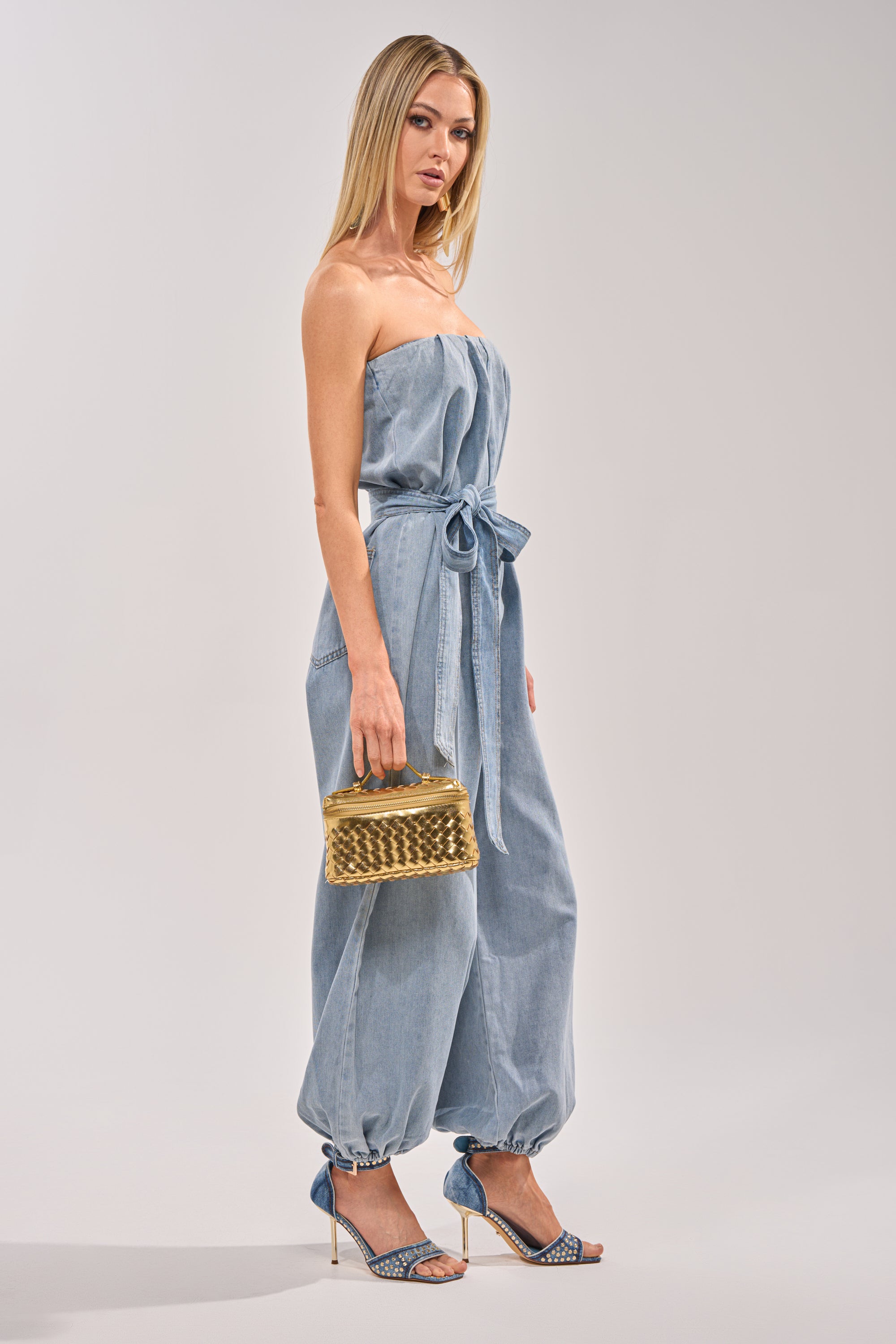 A woman stands against a plain light background wearing the BREEZY DAY DENIM JUMPSUIT with a detachable belt and gathered ankles, paired with blue high heel sandals and holding a small gold woven handbag. 3