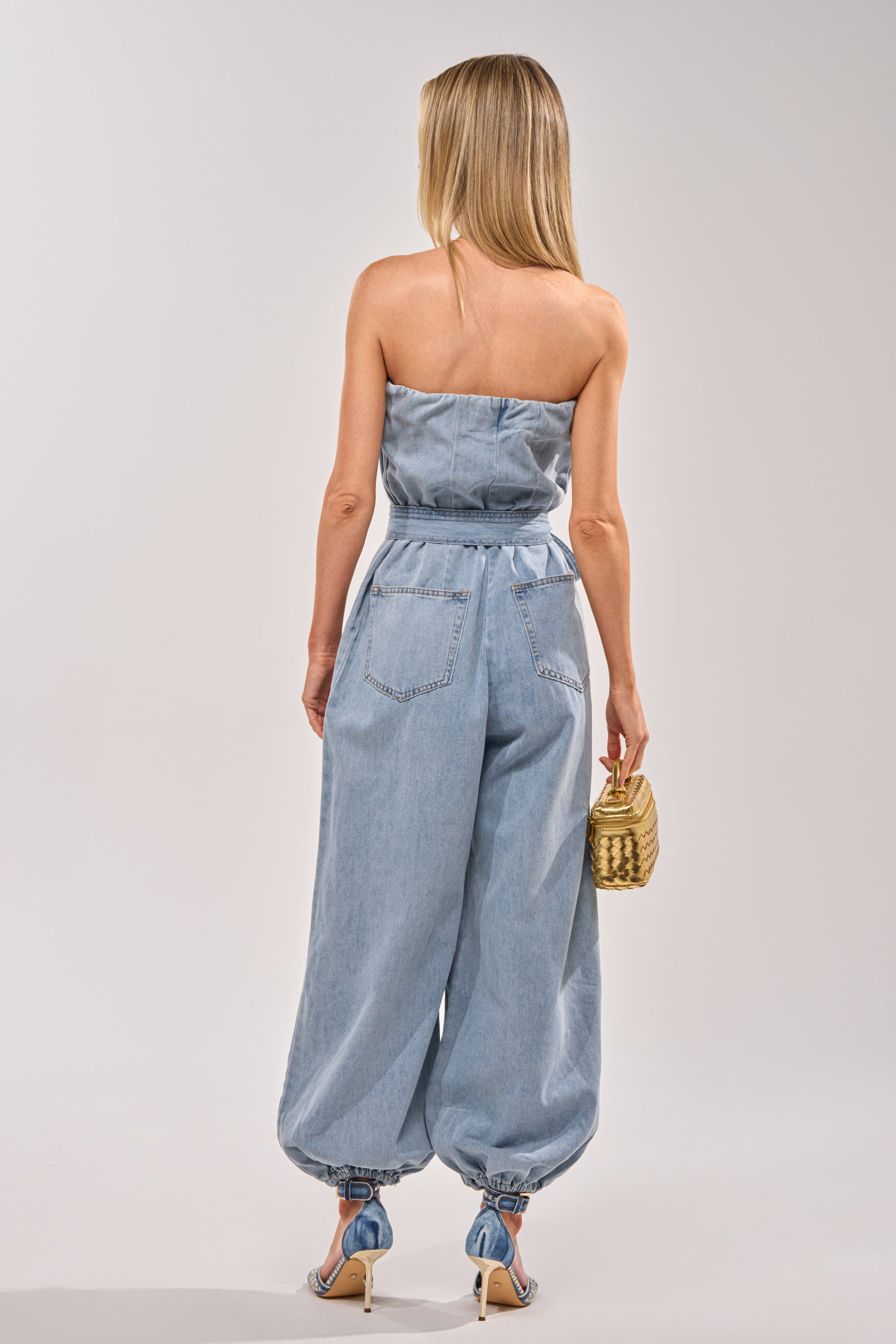 A woman with long blonde hair stands facing away against a plain background, wearing the BREEZY DAY DENIM JUMPSUIT in light-wash denim with cinched ankles, high heels, and holding a small woven handbag in her right hand. 4