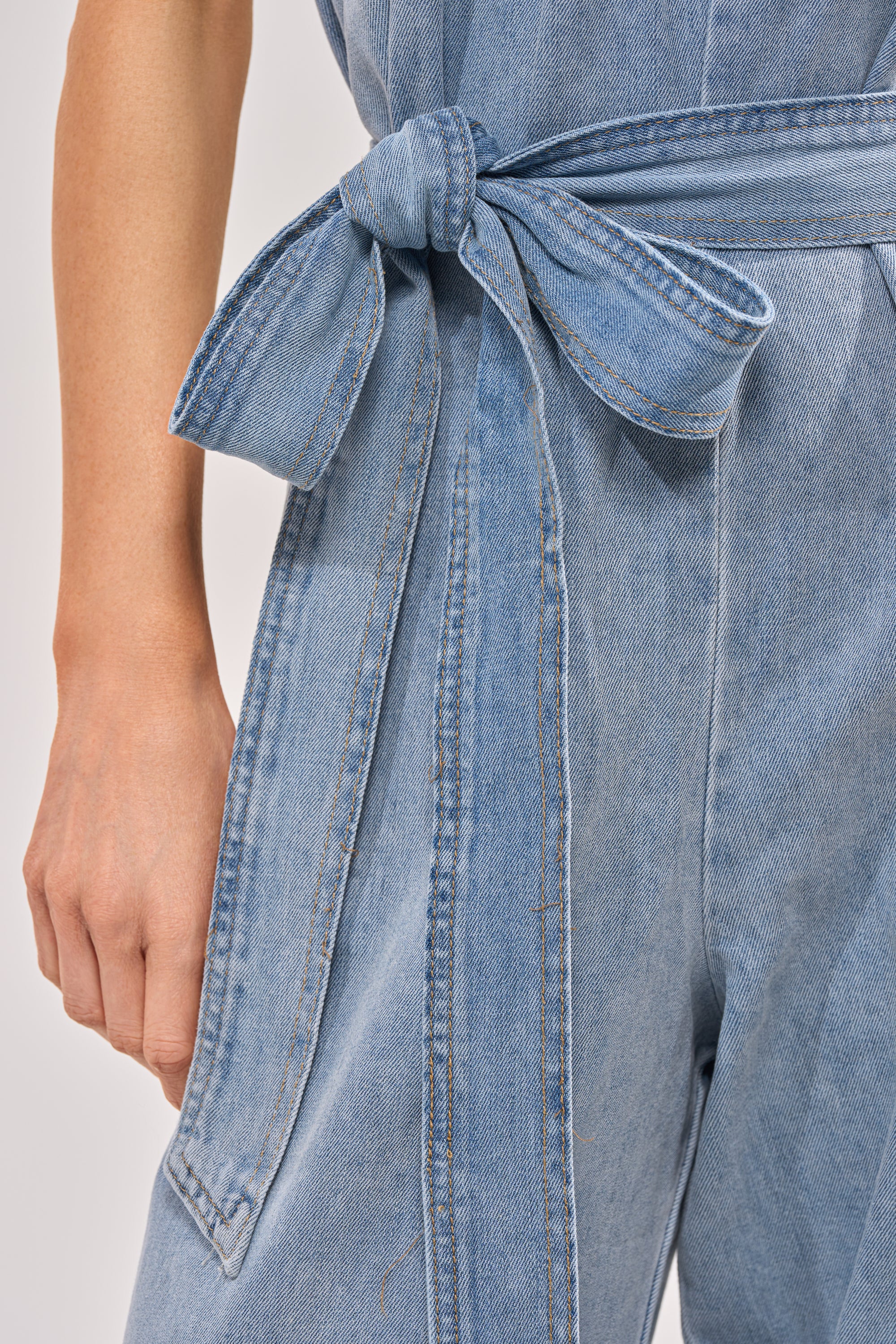 Close-up of a person wearing the BREEZY DAY DENIM JUMPSUIT, featuring light-wash denim and a detachable belt tied in a bow at the waist. Their left arm rests by their side against a plain background. 5