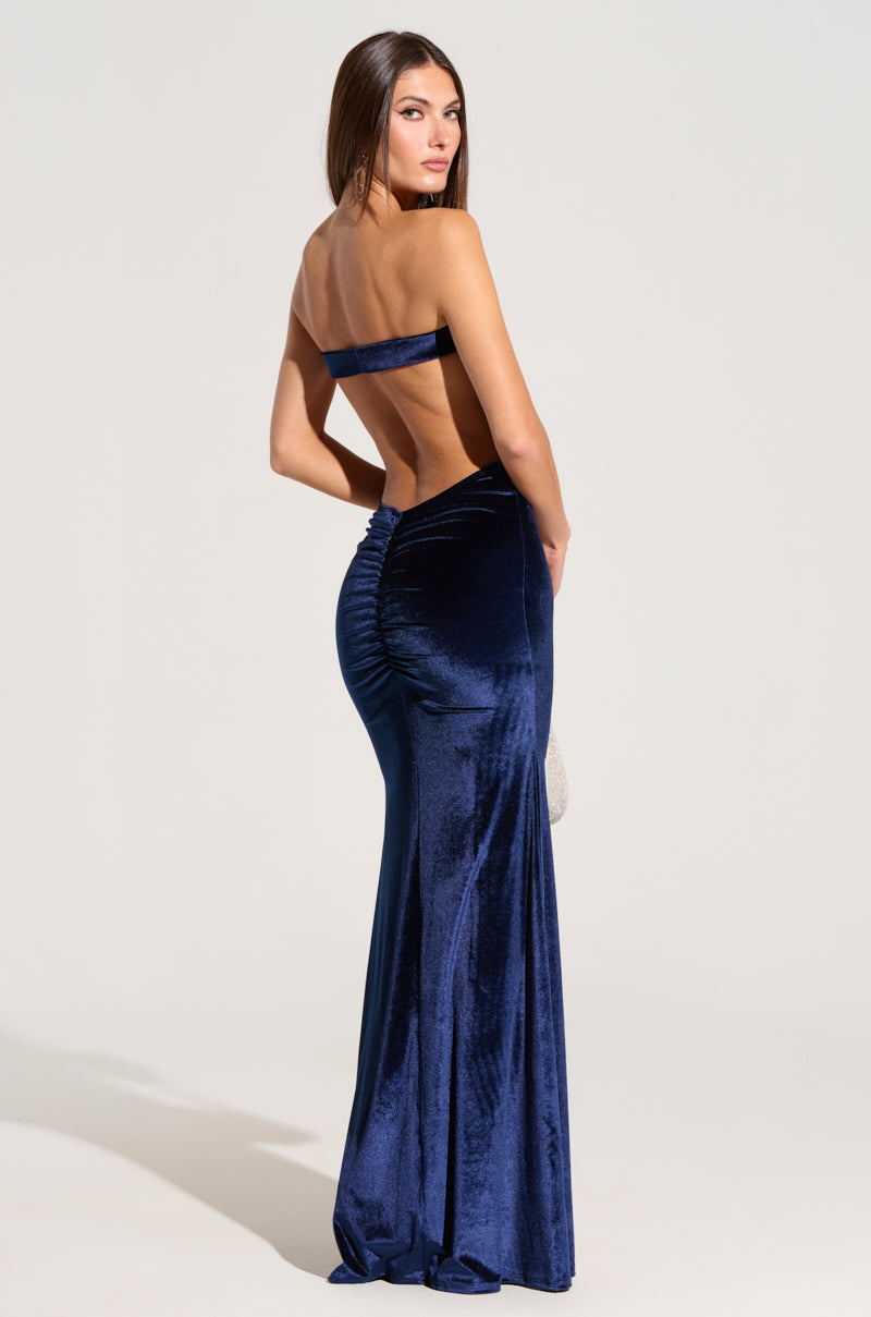 A woman poses with her back turned, wearing the Lost Your Chance Navy Velvet Maxi Dress. This elegant floor-length gown is dark blue and backless, showcasing ruching at the center of the rear. She stands against a plain white background, emphasizing the gown's sophisticated and fitted design.