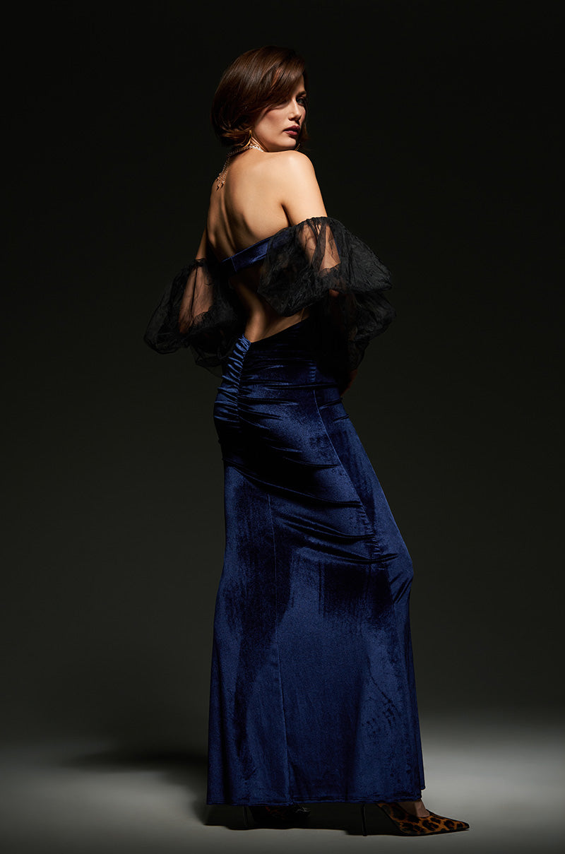 A woman elegantly poses in the LOST YOUR CHANCE Navy Velvet Maxi Dress, featuring flattering ruching and off-shoulder sheer sleeves. She stands against a dark background, highlighting the dress's elegant draping and texture of this glamorous backless gown.