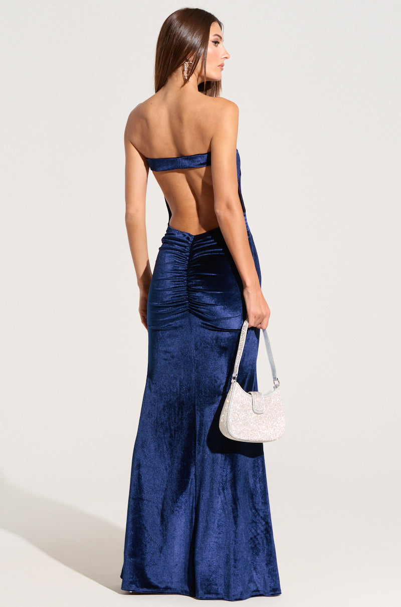 A woman is seen in the LOST YOUR CHANCE Navy Velvet Maxi Dress, a strapless, body-hugging gown with ruching near the waist and a smooth, flowing skirt. She stands facing away, holding a sparkly silver handbag in her right hand as her long brown hair cascades down her back.