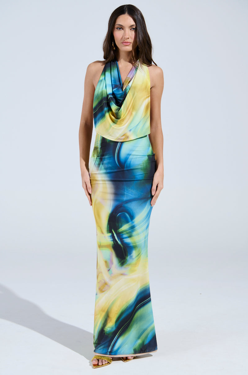 A woman with long, dark hair poses against a neutral background in the NOT IN THE MOOD PRINTED MAXI DRESS, featuring a vibrant swirling pattern of blue, yellow, and green hues. The silky maxi dress has an open-back design and cowl neckline.
