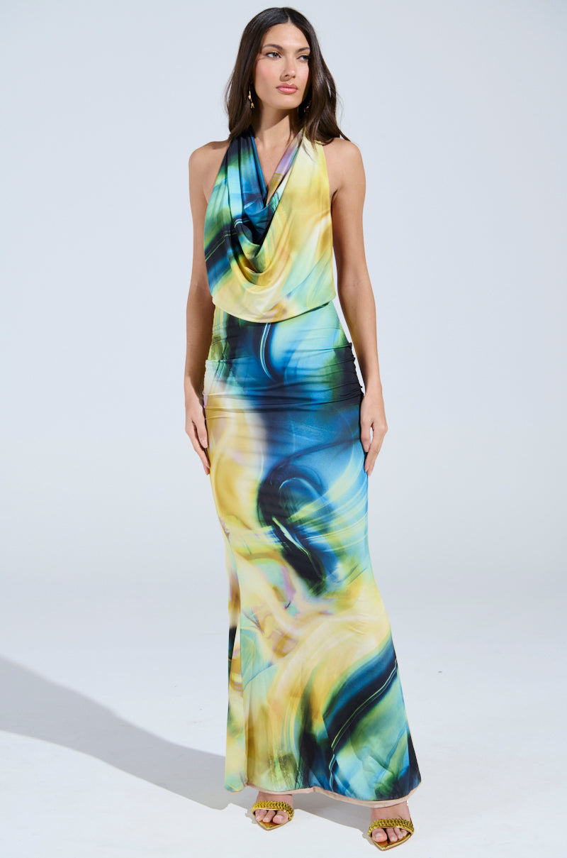 A woman with long dark hair wears the NOT IN THE MOOD PRINTED MAXI DRESS, a silky sleeveless maxi with blue, yellow, and green swirling patterns, featuring an open-back design and draped neckline. She pairs it with open-toed yellow heels against a plain light background.