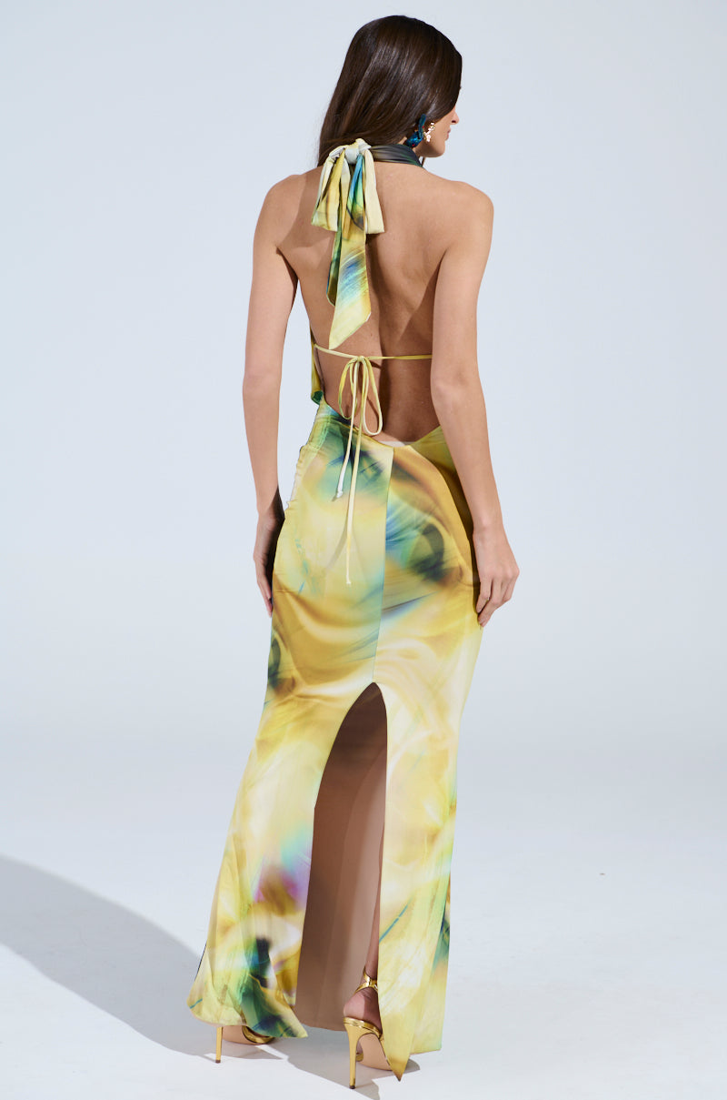 The woman stuns in the NOT IN THE MOOD PRINTED MAXI DRESS, a yellow and green halter-style with an open-back design. This silky maxi dress features a thigh-high slit and swirling pattern, perfectly complemented by her long brown hair, dangling earrings, and high-heeled sandals.