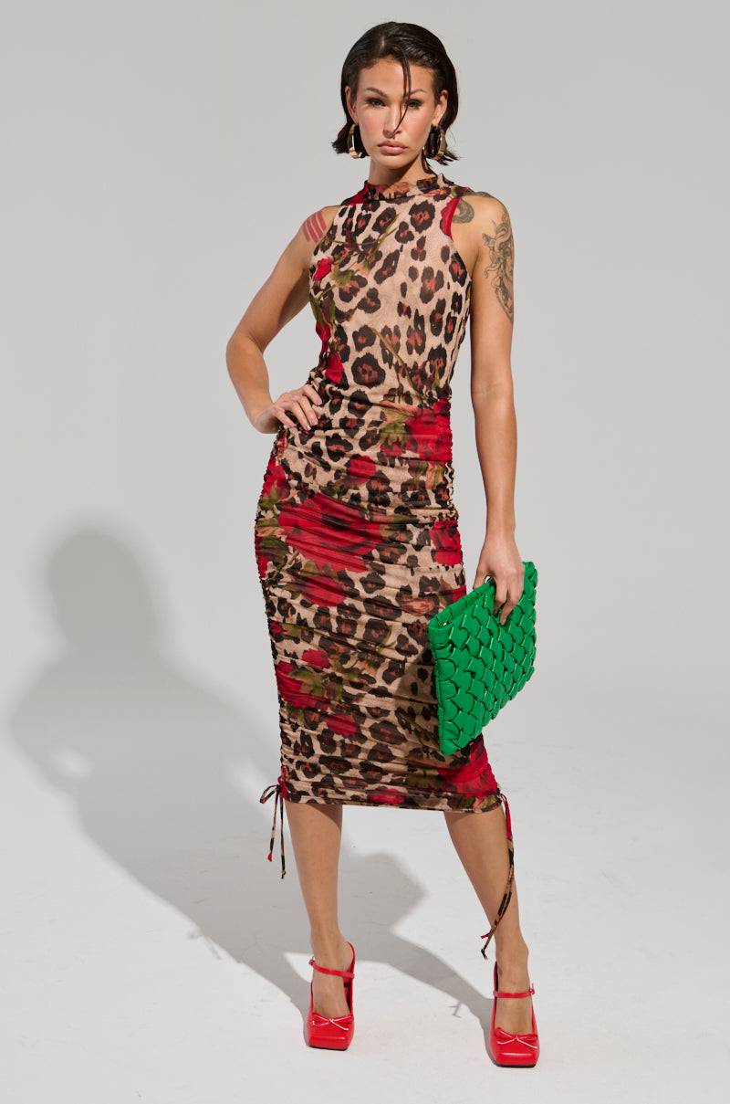The individual exudes confidence in the KALILAH PRINTED MESH MIDI DRESS, a figure-hugging garment that elegantly blends floral and leopard patterns. They accessorize with an oversized green woven clutch and red high heels, their short dark hair complementing the minimalistic gray backdrop.