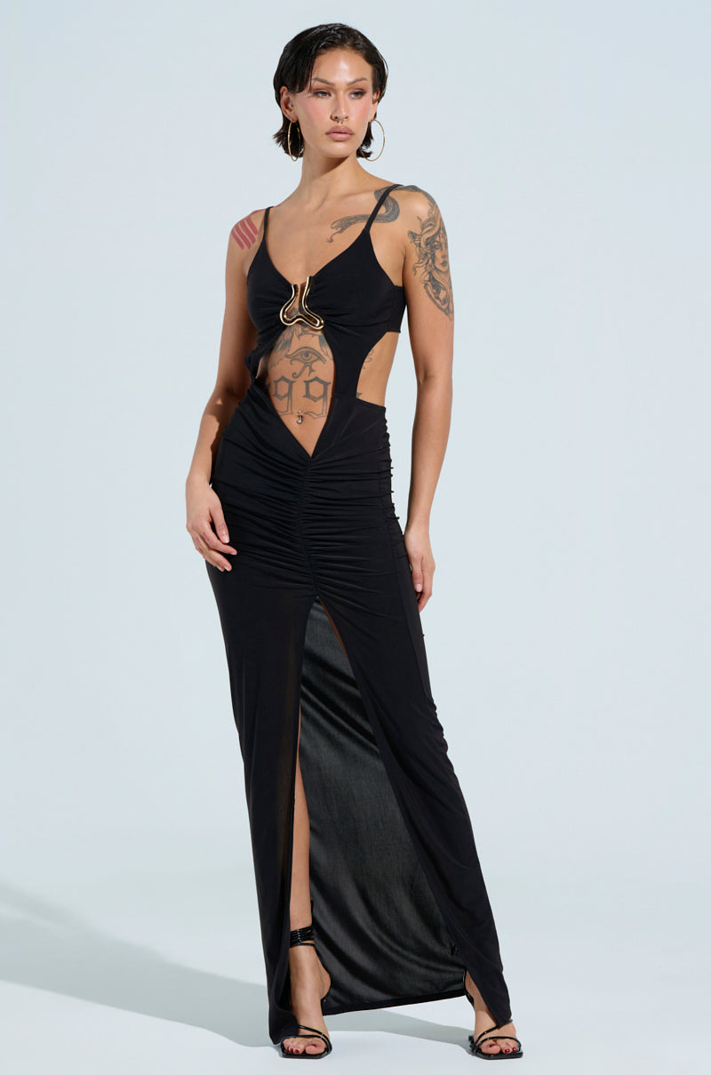 A person wearing the LAY IT LOW SLINKY MAXI DRESS in stylish black, featuring cut-out details, a slit, and a gold chest embellishment, stands confidently. They have tattoos on their arms and wear black high-heeled sandals.