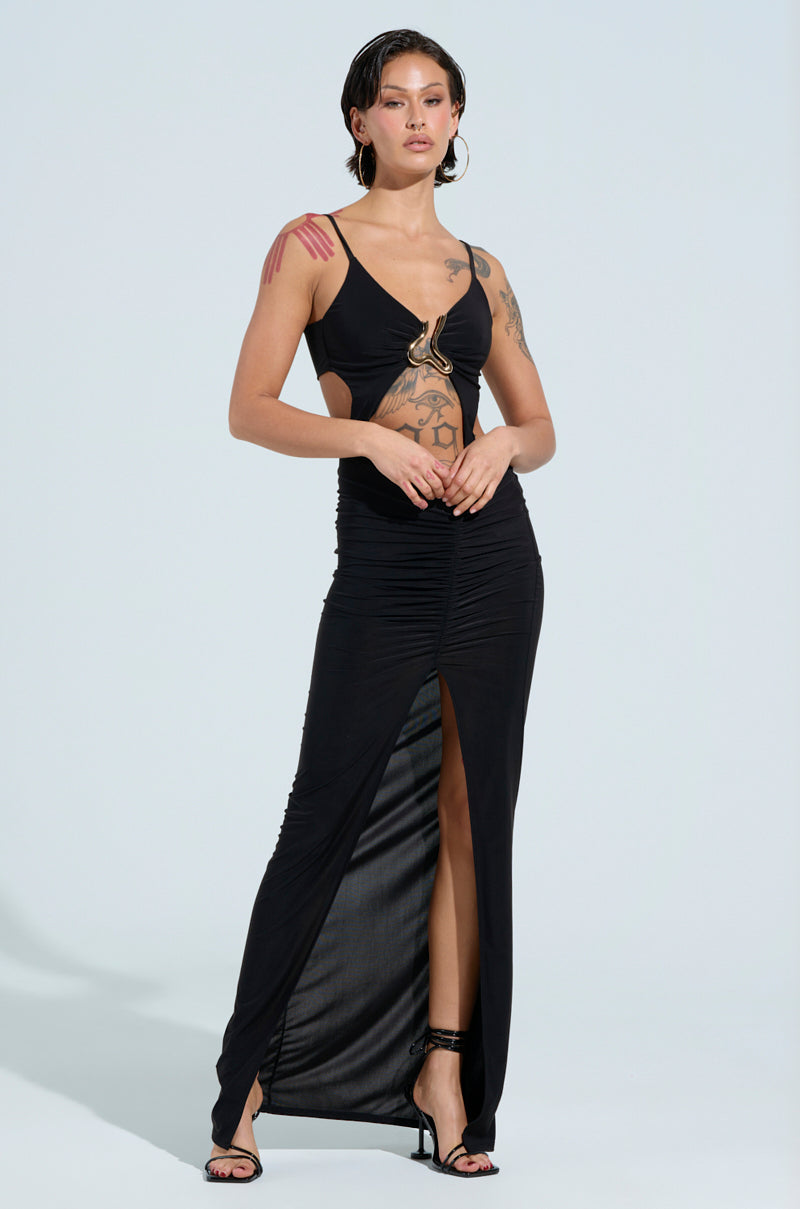 Wearing the LAY IT LOW SLINKY MAXI DRESS with cut-out details and a thigh-high slit, the person exudes confidence. Tattoos grace their arms and chest, paired with black high heels and short styled hair, creating a striking contrast against the neutral light background.