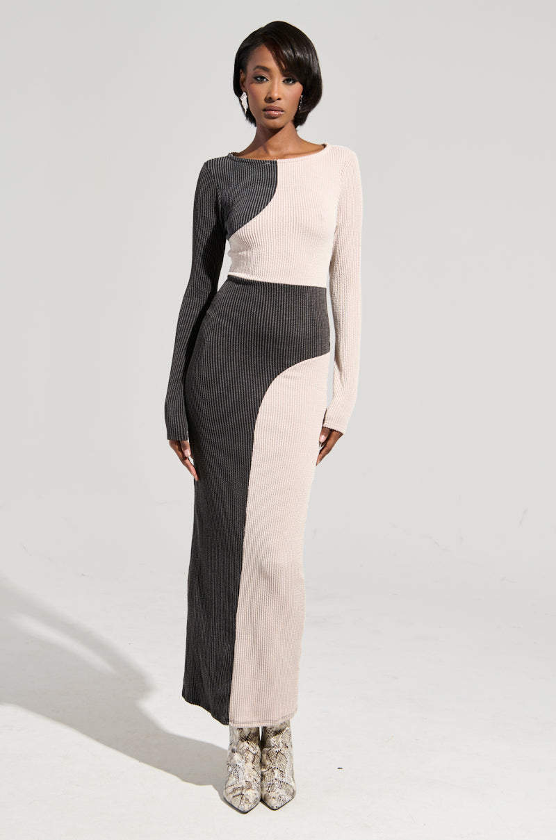 A person stands against a plain background, wearing the TIME TO BALANCE MAXI DRESS—a long-sleeved, bodycon silhouette maxi dress with large asymmetrical black and light beige panels—paired with short hair and snake-patterned boots.