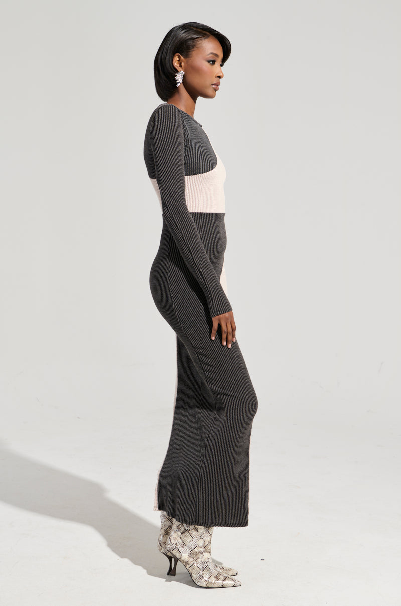 A woman stands in profile against a light background, highlighting the "TIME TO BALANCE MAXI DRESS," a black and beige bodycon silhouette maxi dress with long sleeves. Her look is completed by white snakeskin patterned boots. With short, dark hair and earrings, she exudes modern elegance with flair.
