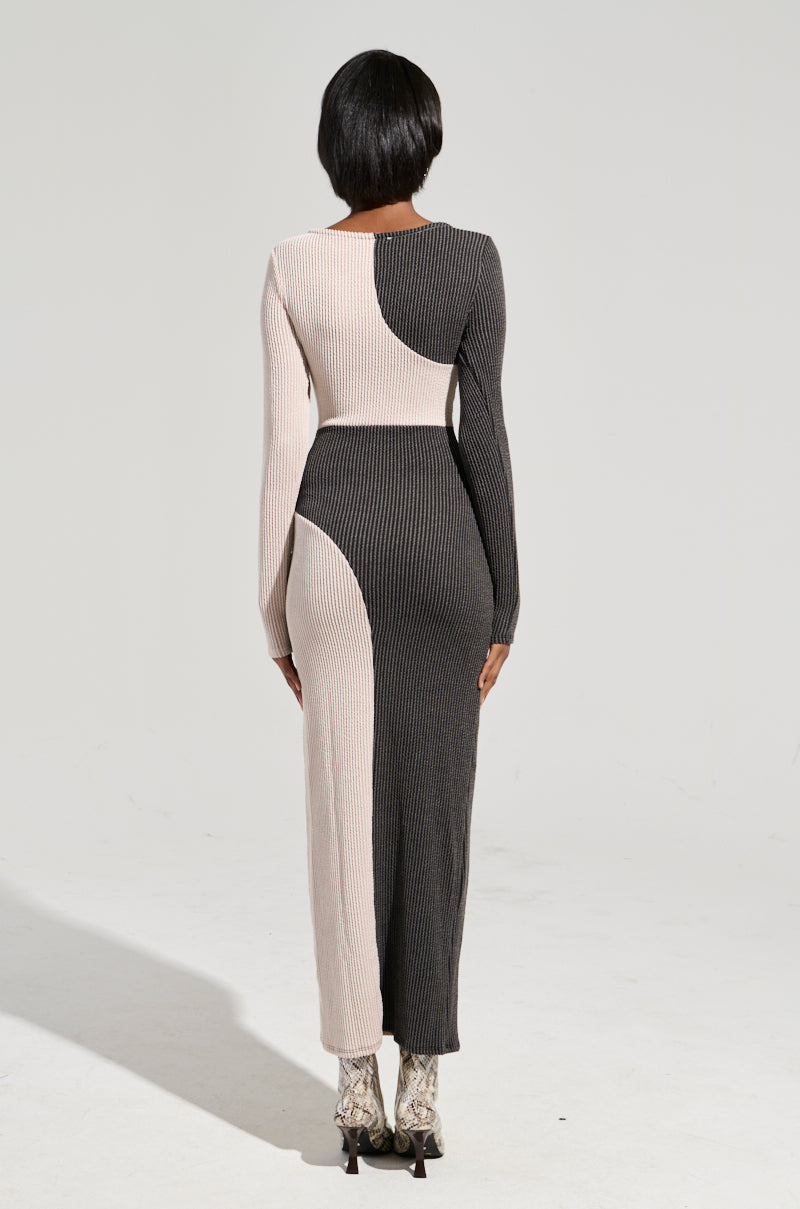A person with short black hair is wearing the "TIME TO BALANCE MAXI DRESS," a long-sleeved, full-length knit maxi dress adorned with geometric patterns in beige and dark gray. They stand on a plain light-colored floor, sporting high-heeled boots, with a neutral-shaded background behind them.