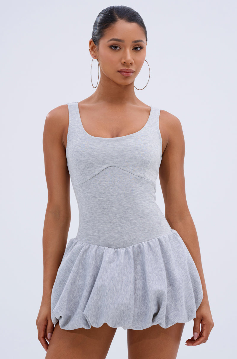 A woman with long hair pulled back, wearing large hoop earrings and a sleeveless GINA FRENCH TERRY MINI DRESS featuring a fitted bodice and bubble hemline, stands against a plain light background.