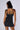 A person with shoulder-length dark hair, wearing the sleeveless, body-hugging WORK IT GIRL KNIT UTILITY DRESS stands facing away from the camera. The person has various tattoos on their arms, back, and legs and is adorned with large hoop earrings and a necklace with small pendants.