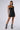 Against a plain white background, a person showcases the "WORK IT GIRL KNIT UTILITY DRESS," a sleeveless, form-fitting black mineral wash mini dress featuring zipper details and front pockets. The bodycon dress is elegantly accessorized with a chunky necklace, earrings, and open-toed ankle boots in an acid-wash style.