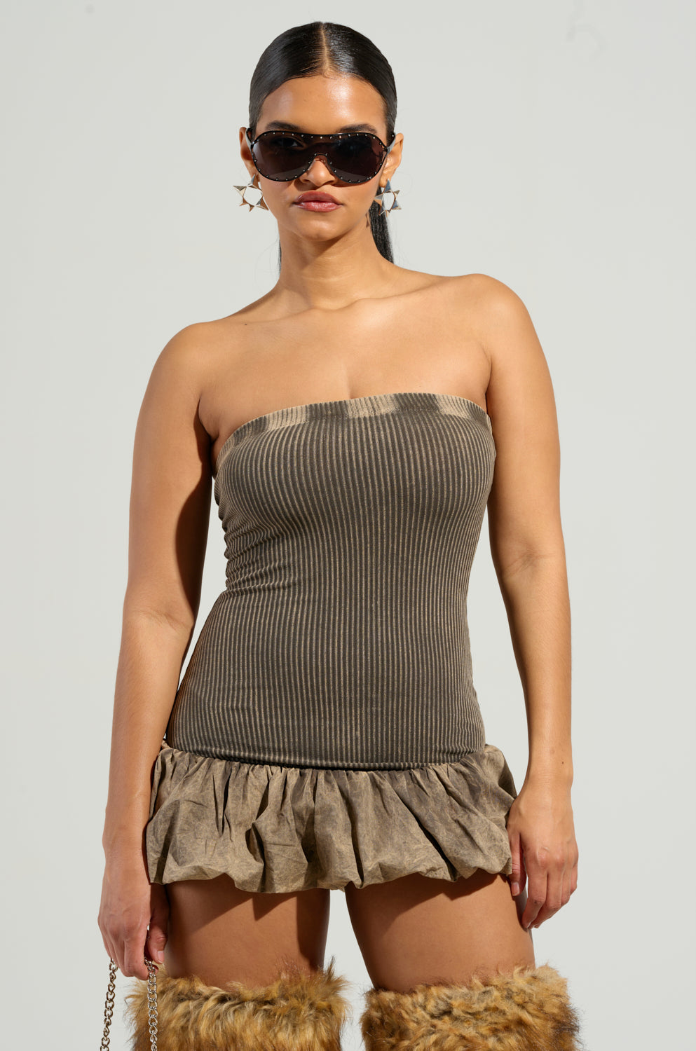 A woman confidently stands in an AKIRA Label bodycon fit, styled with the STAY WITH ME MINI DRESS and furry knee-high boots. She accessorizes with large sunglasses and star-shaped earrings, her hair elegantly pulled into a sleek ponytail.