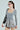 A person with a ponytail wears glasses and large hoop earrings. They are dressed in a GREY GIVE ME A LITTLE MINI DRESS. A black shoulder bag hangs on one arm against a light gray background.