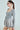 A confident person with a ponytail and glasses stands sideways against a white background, wearing the 