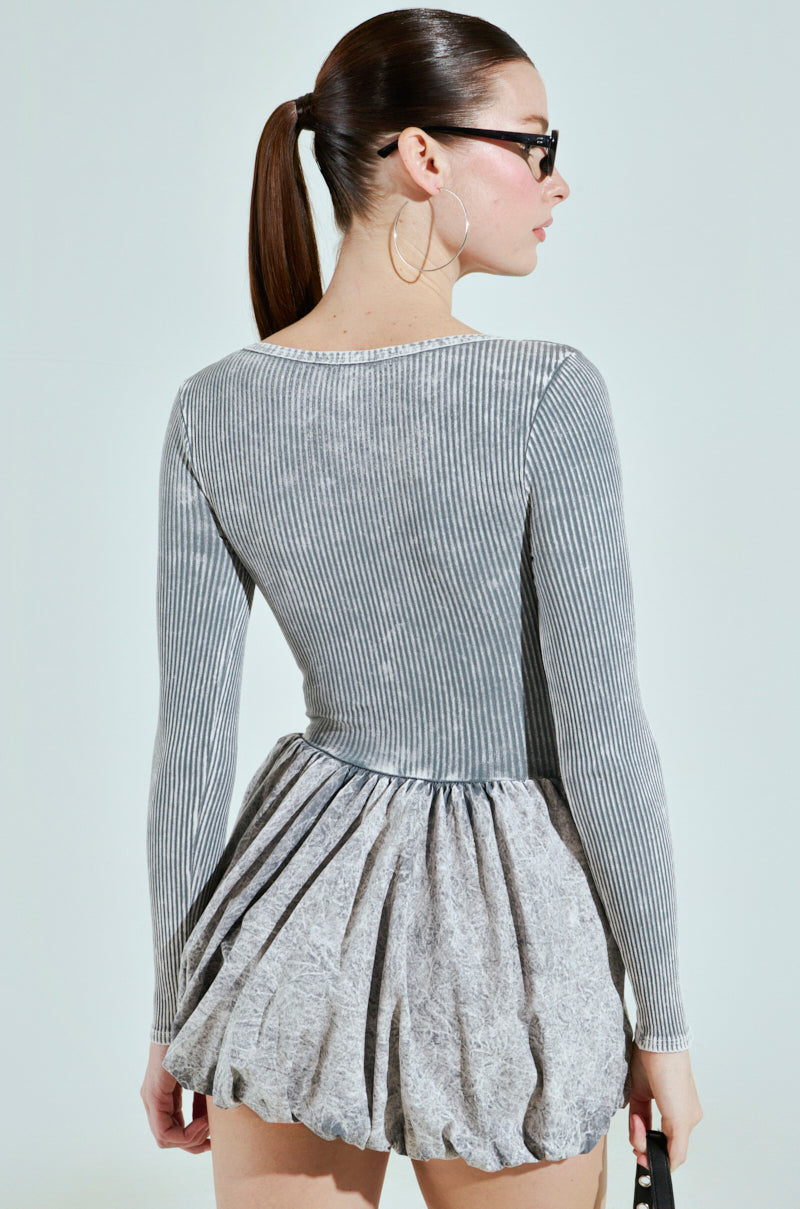 A woman with a ponytail, glasses, and hoop earrings is seen from behind. She wears the "Give Me a Little" mini dress in light gray, holding a black bag against a light gray backdrop.