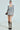 A woman in a ribbed knit light grey GIVE ME A LITTLE MINI DRESS, paired with a voluminous textured skirt and knee-high lace-up boots. She's accessorized with glasses, hoop earrings, and holding a black handbag in a well-lit studio with a neutral background.