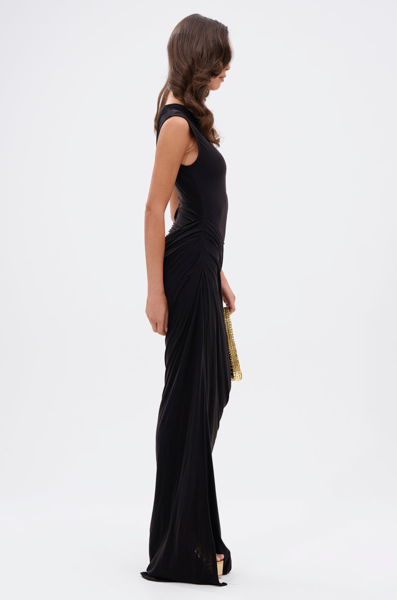 A woman stands in profile against a white background, wearing the ONLY ME CUT-OUT MIDI DRESS IN BLACK with a gold clutch, black high heels, and her long hair styled in loose waves.