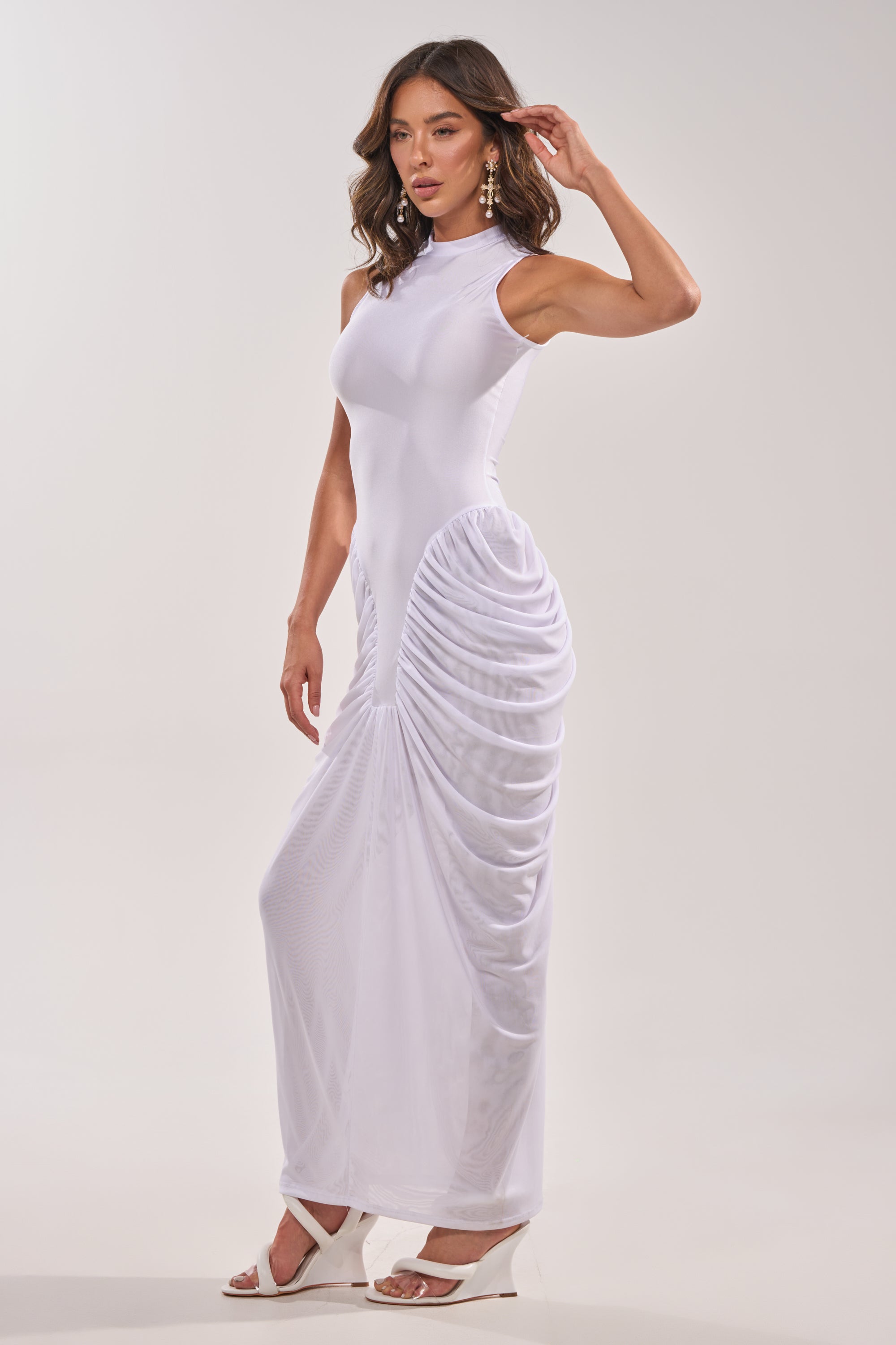 A woman models the SECOND DATE BODYCON SHINE MAXI DRESS IN WHITE, a sleeveless, high-neck ruched maxi with hip detailing. She pairs it with white heeled sandals and statement earrings against a plain light background. 1