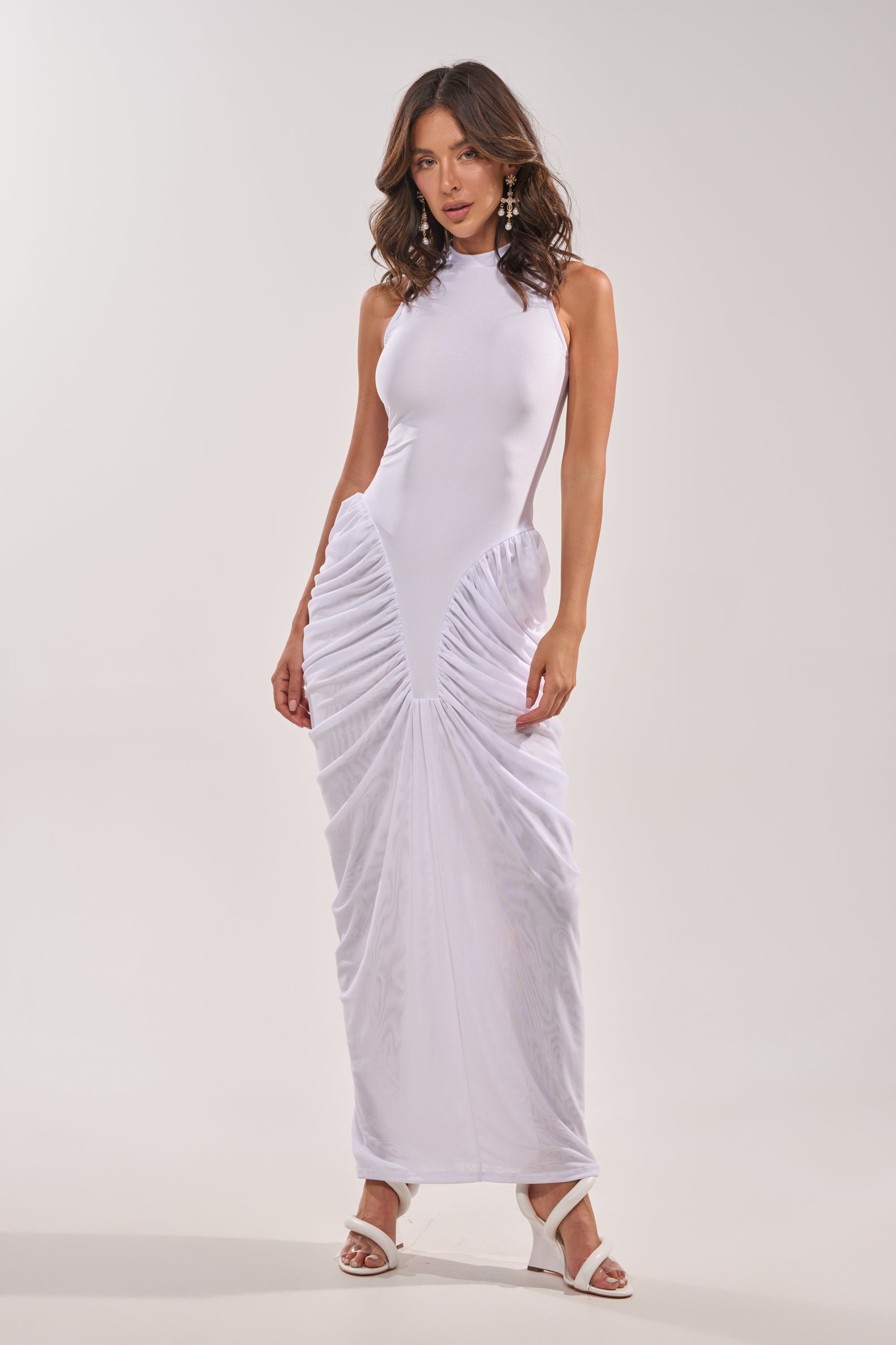 A woman wears the SECOND DATE BODYCON SHINE MAXI DRESS IN WHITE, a sleeveless, high-neck fitted maxi with ruched skirt detailing, paired with white heeled sandals and statement earrings against a plain light background. 2