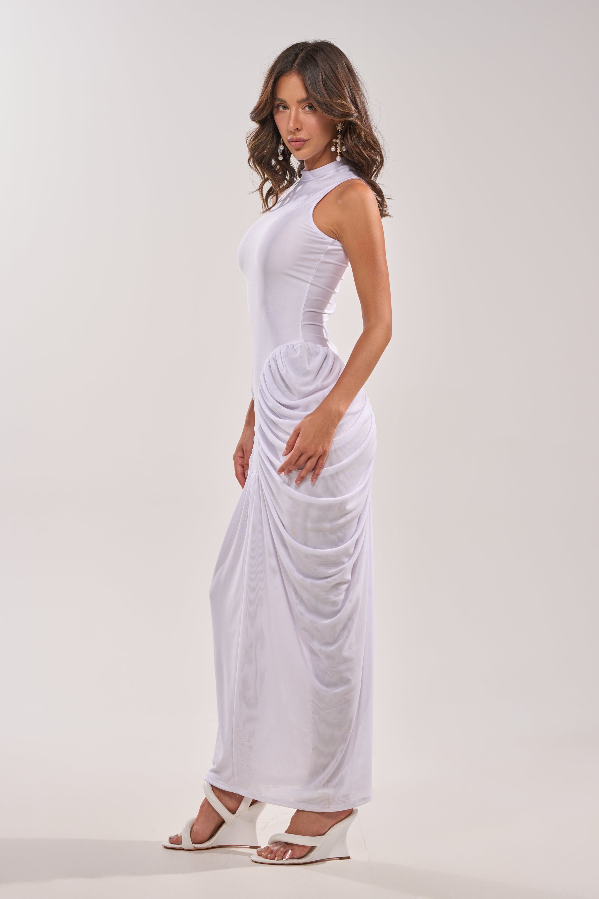 A woman with wavy brown hair wears the SECOND DATE BODYCON SHINE MAXI DRESS IN WHITE and white heeled sandals, standing in profile against a plain, light-colored background. 4