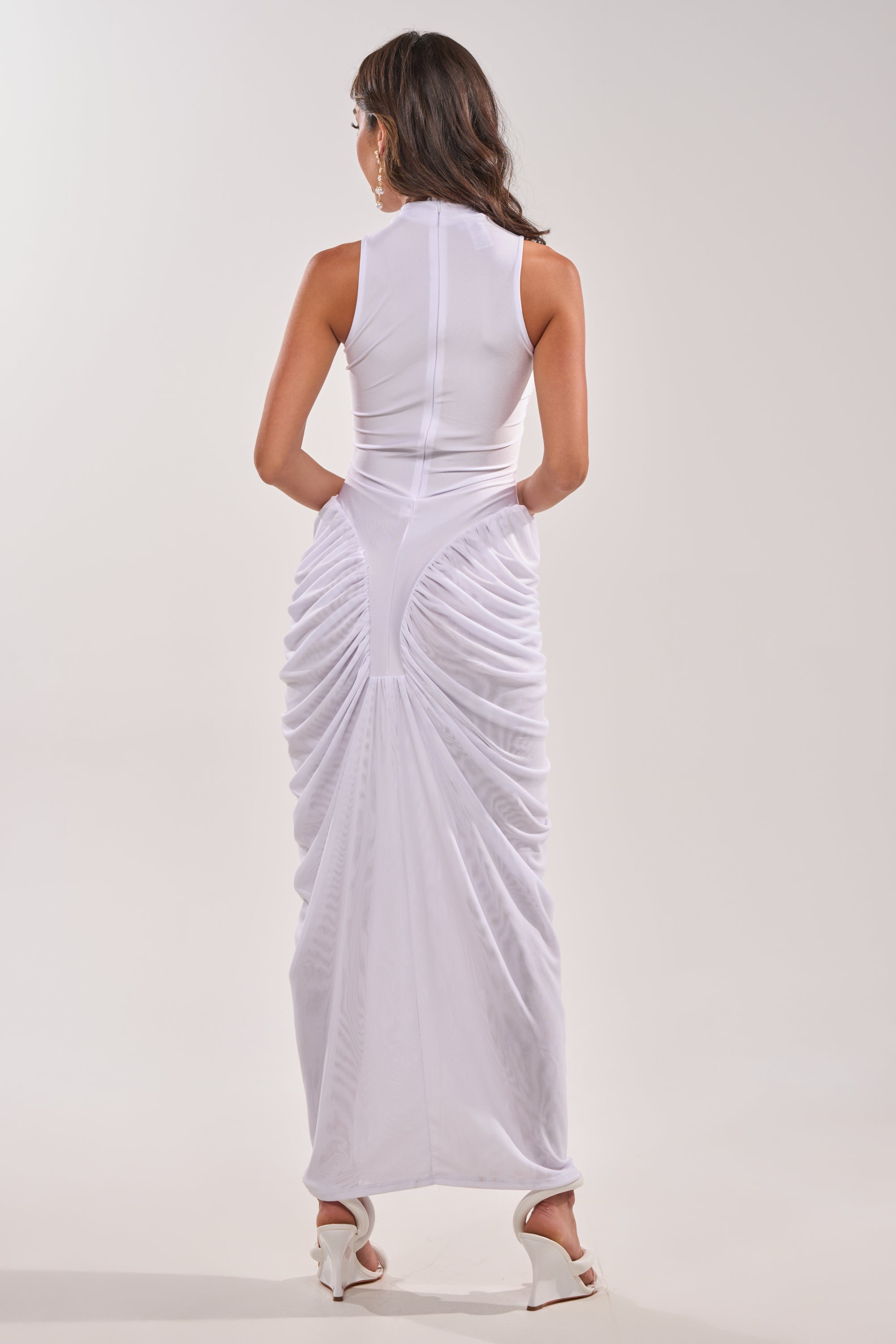 A woman with long, wavy brown hair faces away, wearing the SECOND DATE BODYCON SHINE MAXI DRESS IN WHITE and white heeled sandals against a plain light background. 5