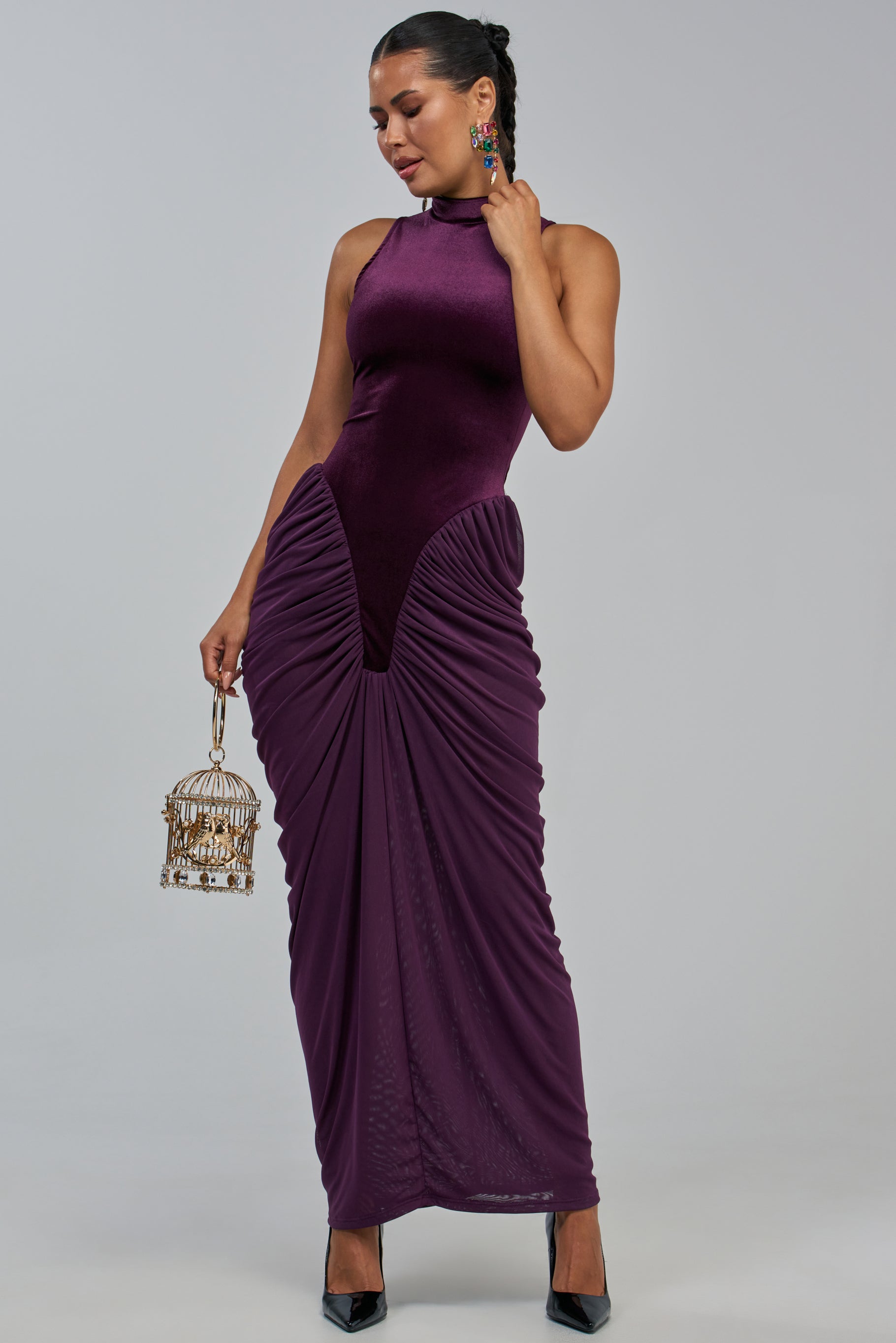 Wearing the SECOND DATE MAXI DRESS IN PLUM, a woman stands against a plain gray background. The sleeveless bodycon maxi dress features ruched detailing, and she completes her look with statement earrings, black heels, and a small, ornate cage-shaped handbag.