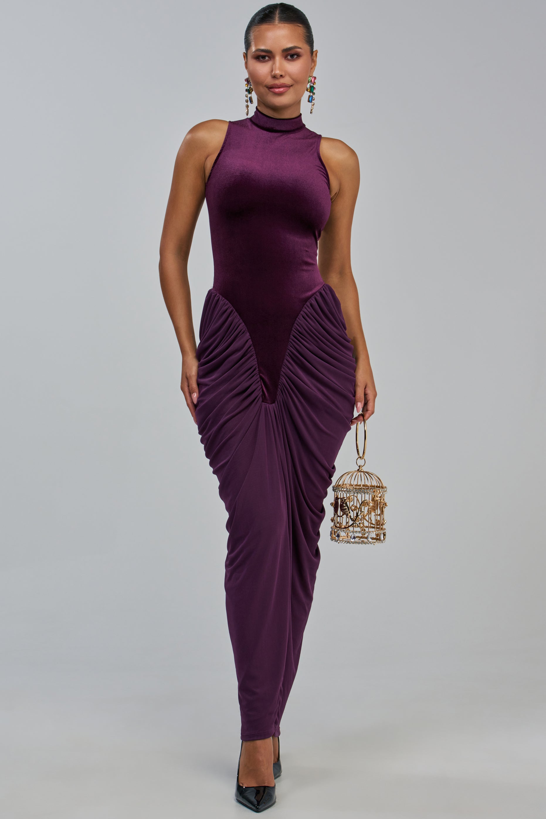 A woman stands against a neutral background wearing the SECOND DATE MAXI DRESS IN PLUM—a sleeveless, high-neck ruched mesh dress with elegant detailing. She holds a small gold cage-shaped purse and wears black heels.