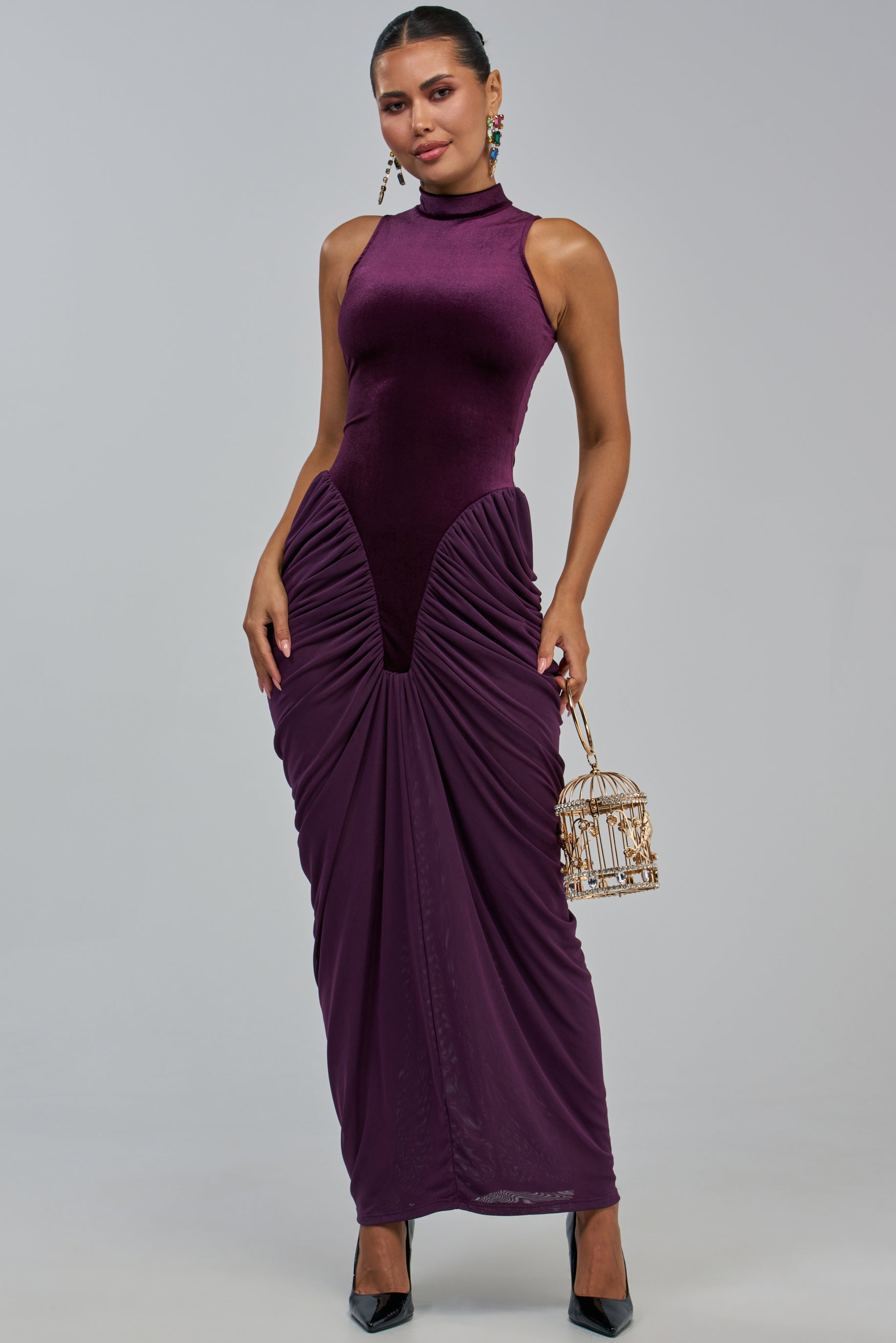 A woman poses confidently against a plain gray background, wearing the SECOND DATE MAXI DRESS IN PLUM—a sleeveless, high-neck bodycon maxi dress featuring ruched detailing. She accessorizes with a small decorative birdcage purse and black heels.