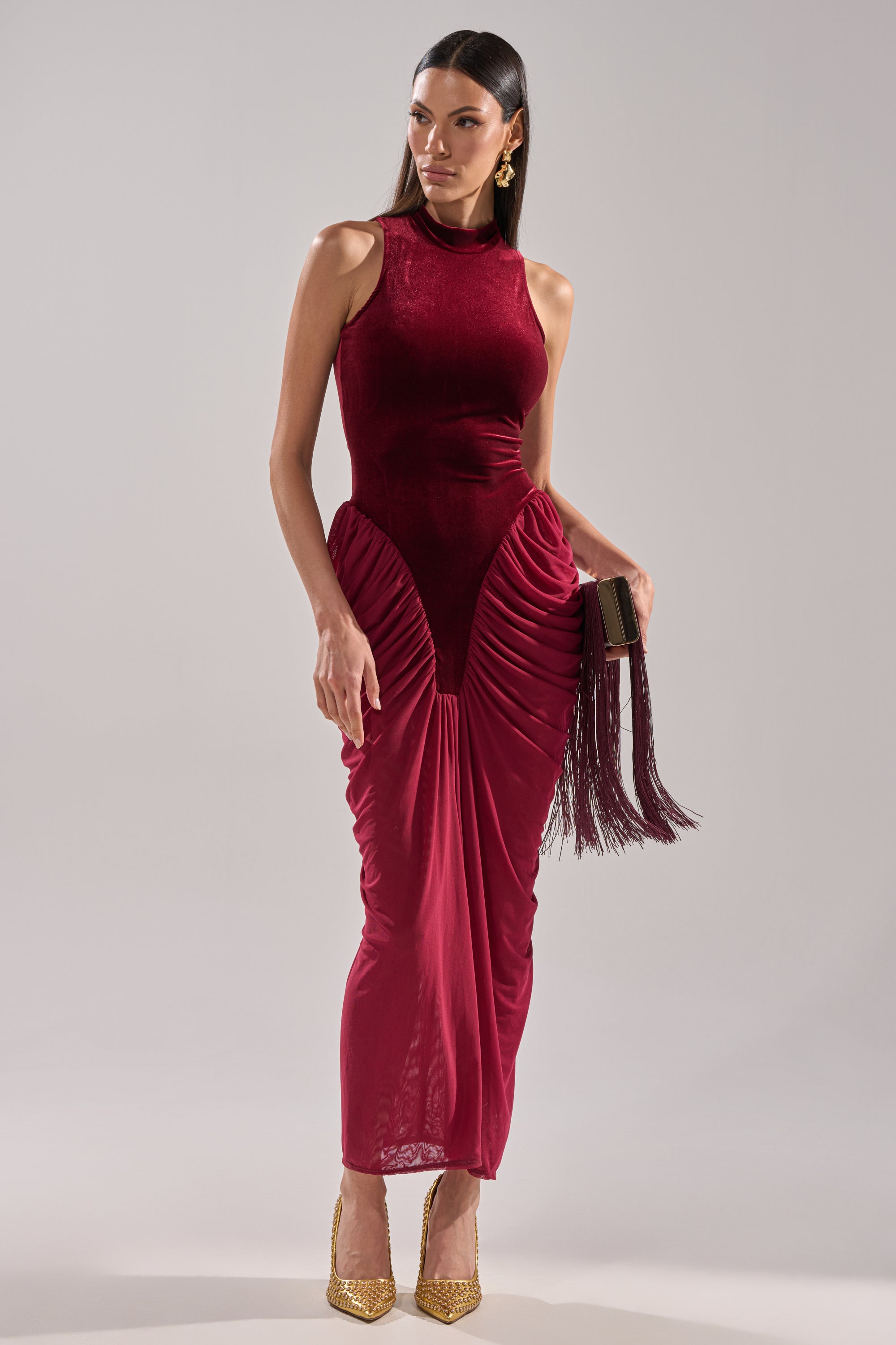 A woman wears the BABY ITS COLD OUTSIDE VELVET RUCHED MAXI DRESS—sleeveless, high-neck, fitted, and burgundy—for a formal event, paired with gold heels, gold earrings, and a black fringed clutch against a plain light background.
