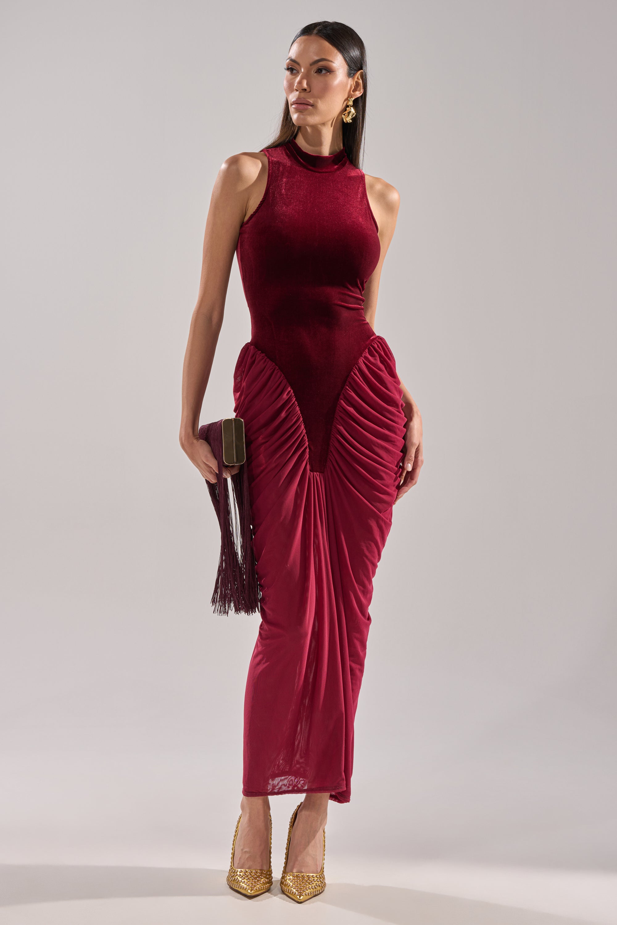 Ideal for formal events, she wears the BABY ITS COLD OUTSIDE VELVET RUCHED MAXI DRESS in burgundy with gold heels and earrings, holding a matching tassel clutch against a plain light background.