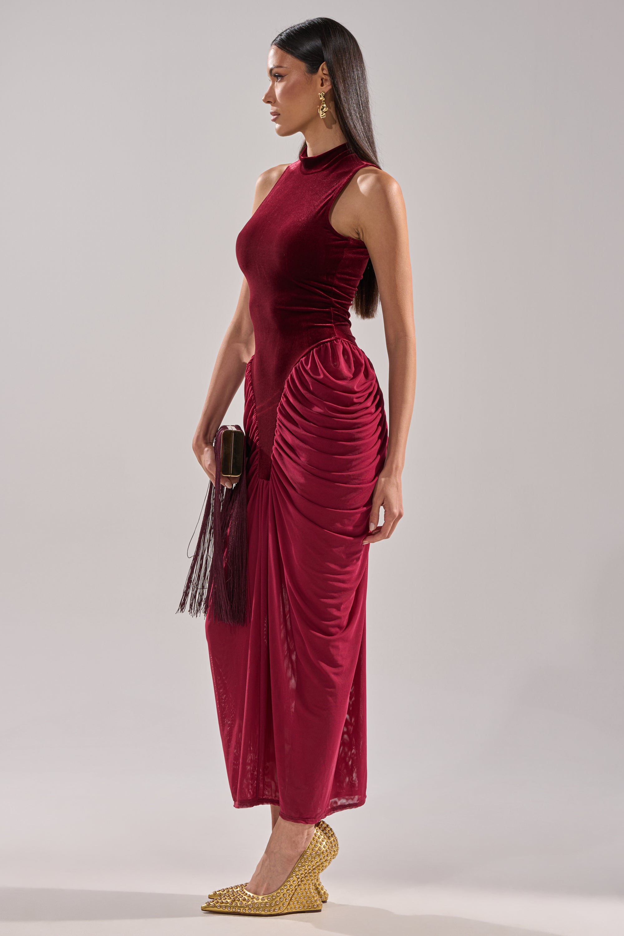 A woman stands in profile wearing the BABY ITS COLD OUTSIDE VELVET RUCHED MAXI DRESS in deep red, paired with gold earrings, gold pointed heels, and a black fringed clutch—an elegant formal look set against a plain gray background.