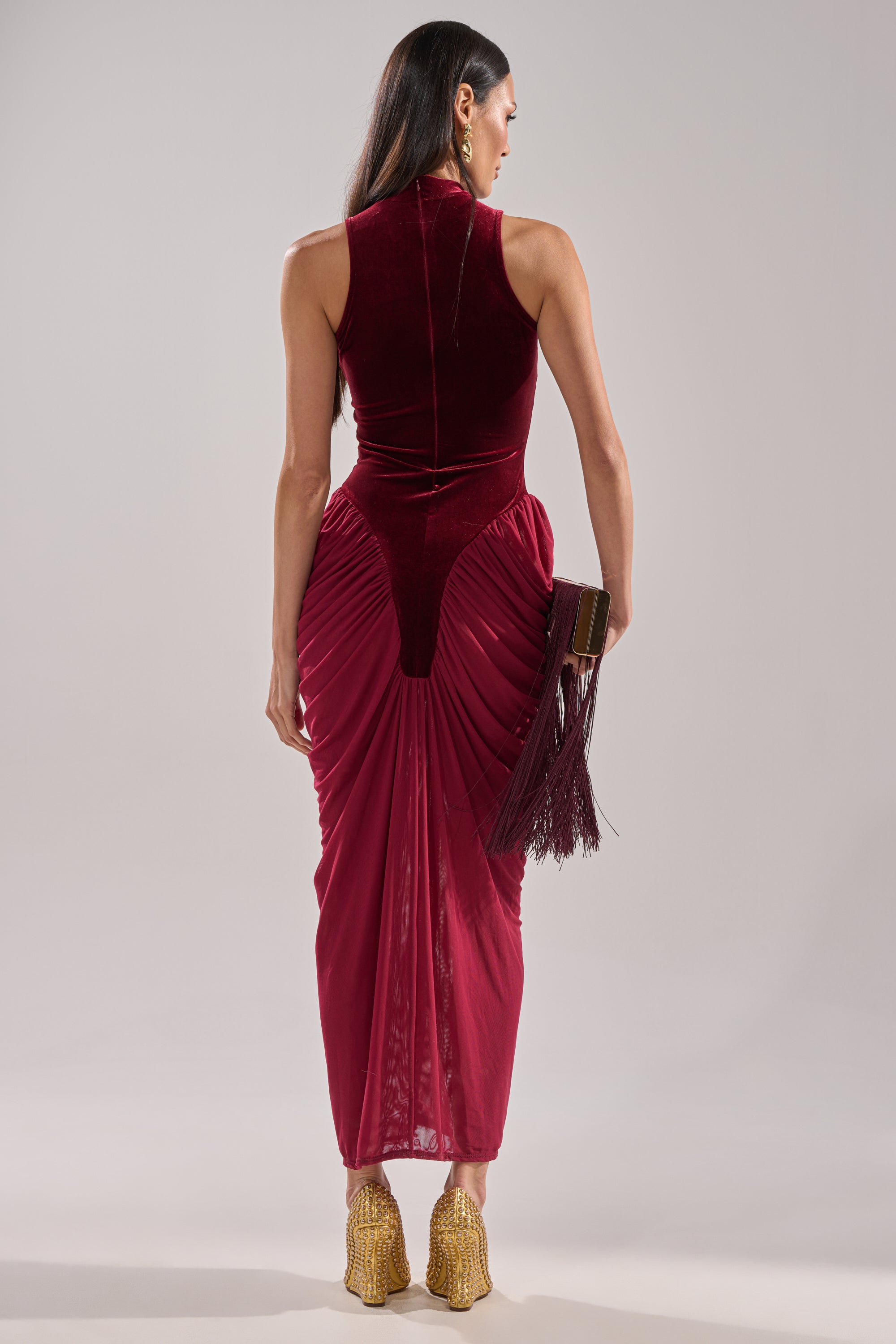 A person with long dark hair wears the BABY ITS COLD OUTSIDE VELVET RUCHED MAXI DRESS, a sleeveless, fitted burgundy velvet maxi with gathered hip details. They hold a fringed clutch and wear gold heels against a plain light background.