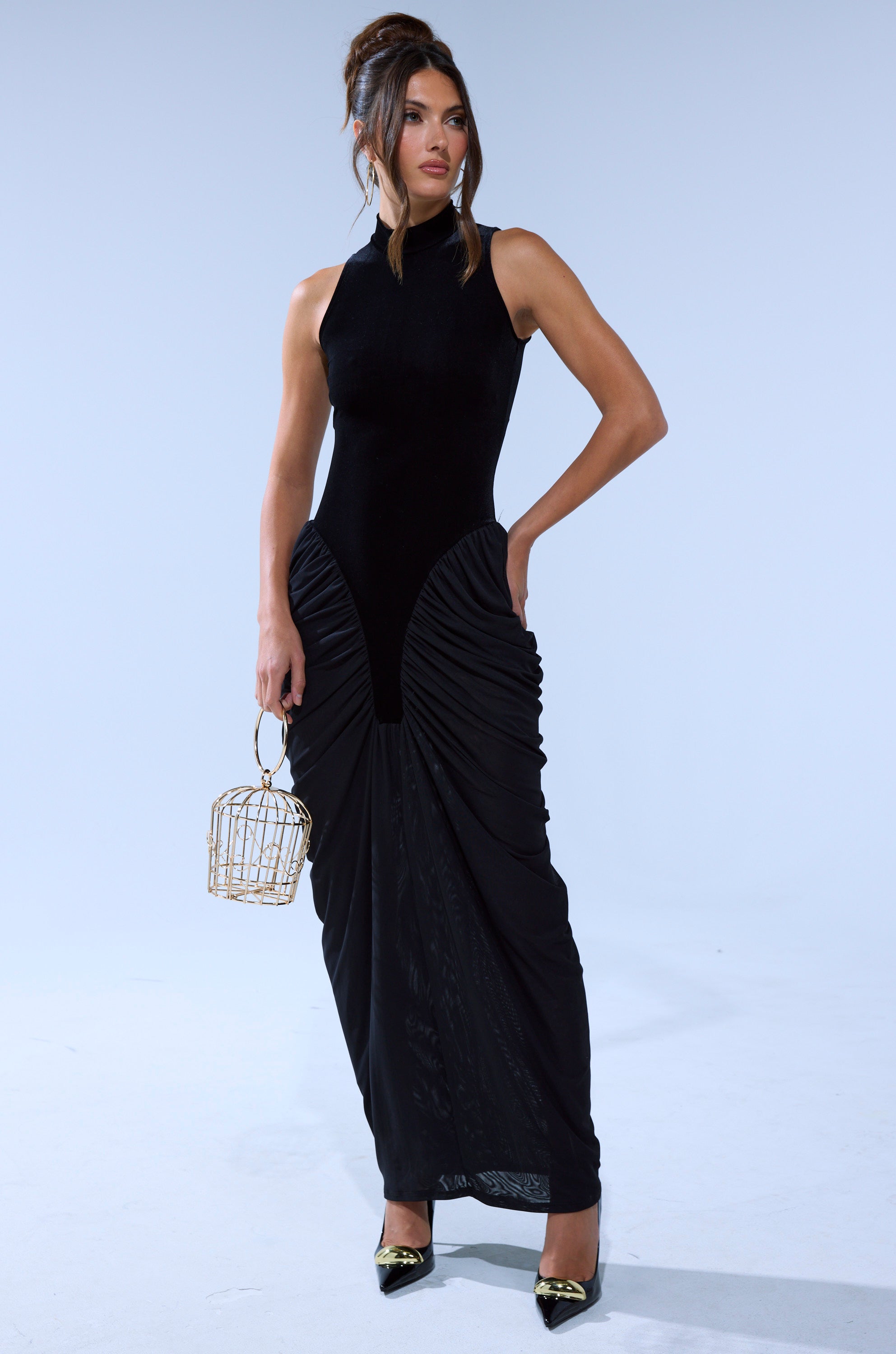 A woman stands against a light background in the OUT FOR THE NIGHT MOCK NECK MAXI DRESS, featuring ruched detailing. She pairs it with black pointed heels, a gold cage handbag, and wears her hair in an elegant updo.