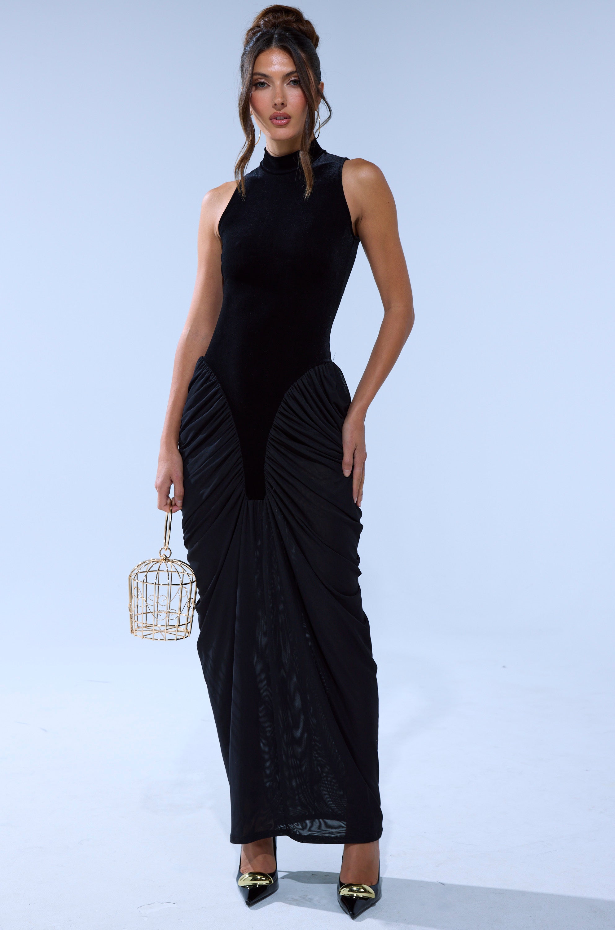 A woman wears the OUT FOR THE NIGHT MOCK NECK MAXI DRESS in black with ruched detailing, stands against a plain background, holding a gold cage-shaped bag and wearing black pointed-toe heels with gold accents.