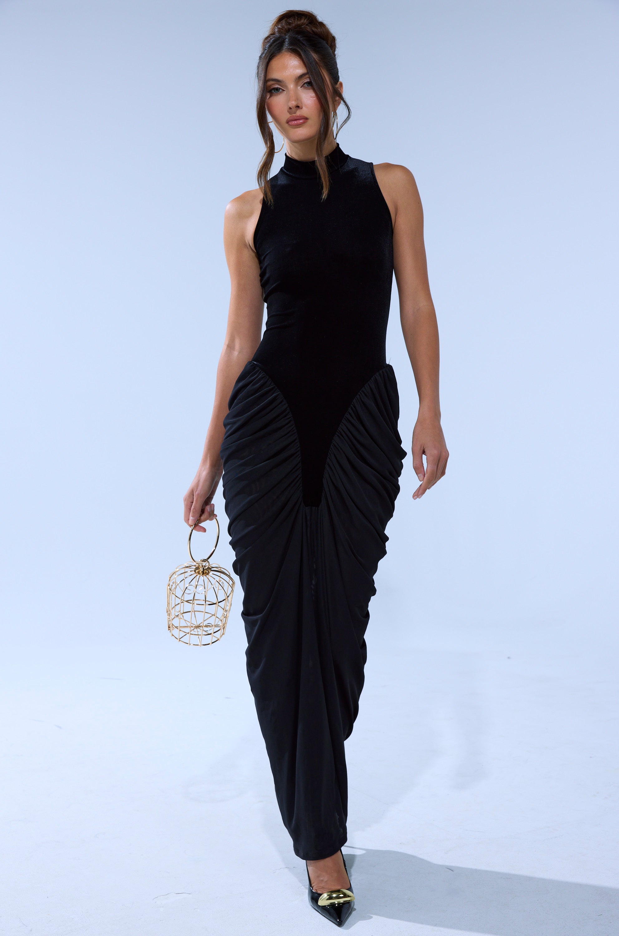 A woman wears the OUT FOR THE NIGHT MOCK NECK MAXI DRESS—black, sleeveless with a high-neck and ruched skirt—styled with black pointed heels and a gold cage handbag, showcasing a chic look against a plain background.
