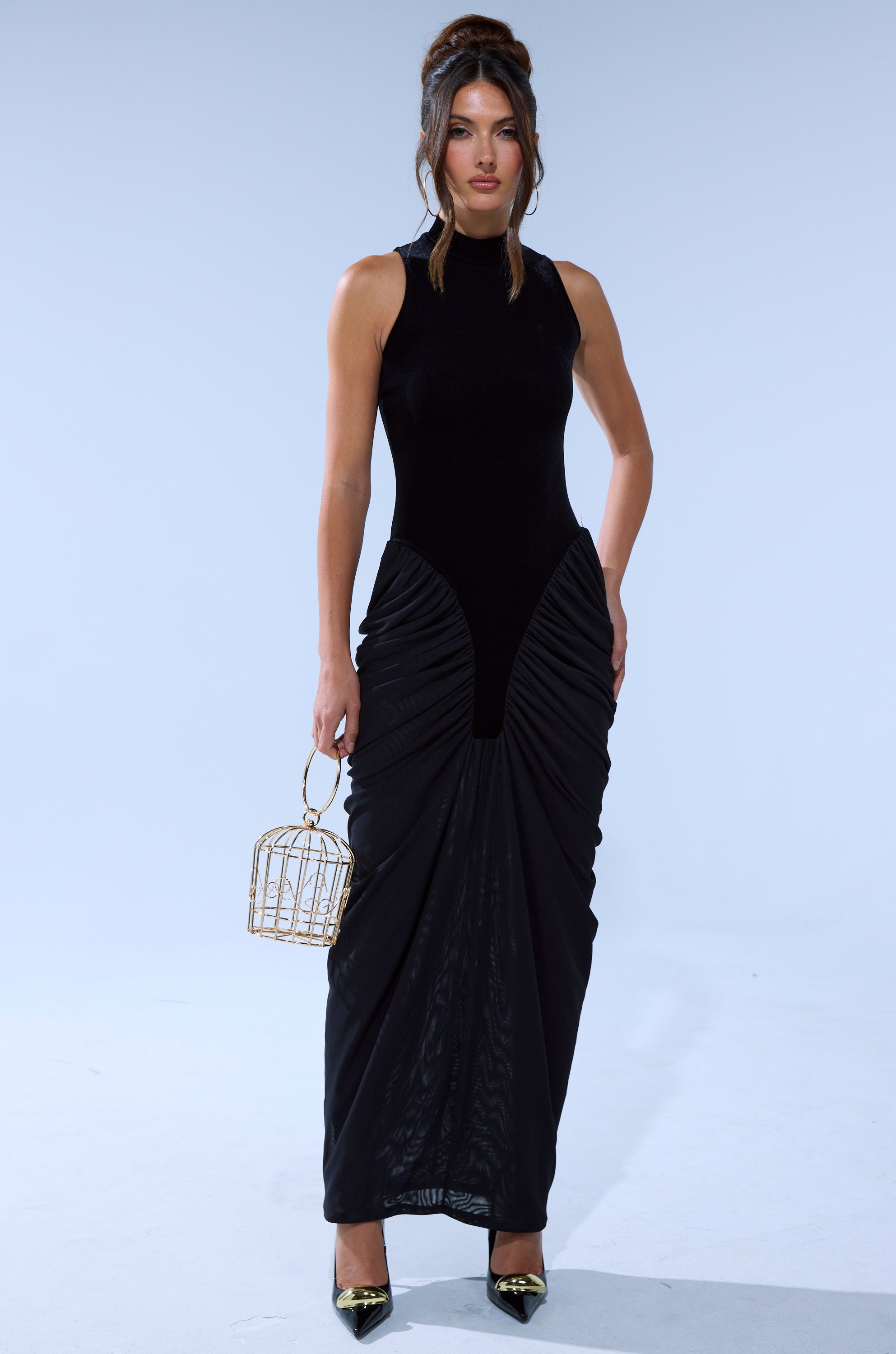 Wearing the OUT FOR THE NIGHT MOCK NECK MAXI DRESS, a woman with an updo and loose strands carries a gold cage purse and wears black pointed heels, creating a sleek, elegant style.