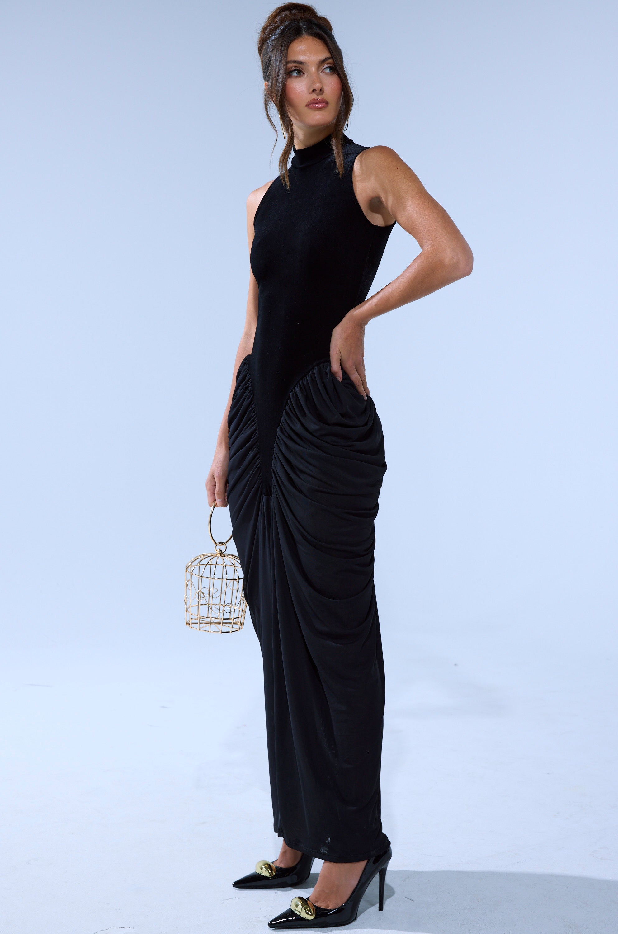 A woman wears the OUT FOR THE NIGHT MOCK NECK MAXI DRESS, a black sleeveless maxi with a high neck and ruched sides, paired with a gold cage handbag and black heels featuring gold accents, against a plain backdrop.
