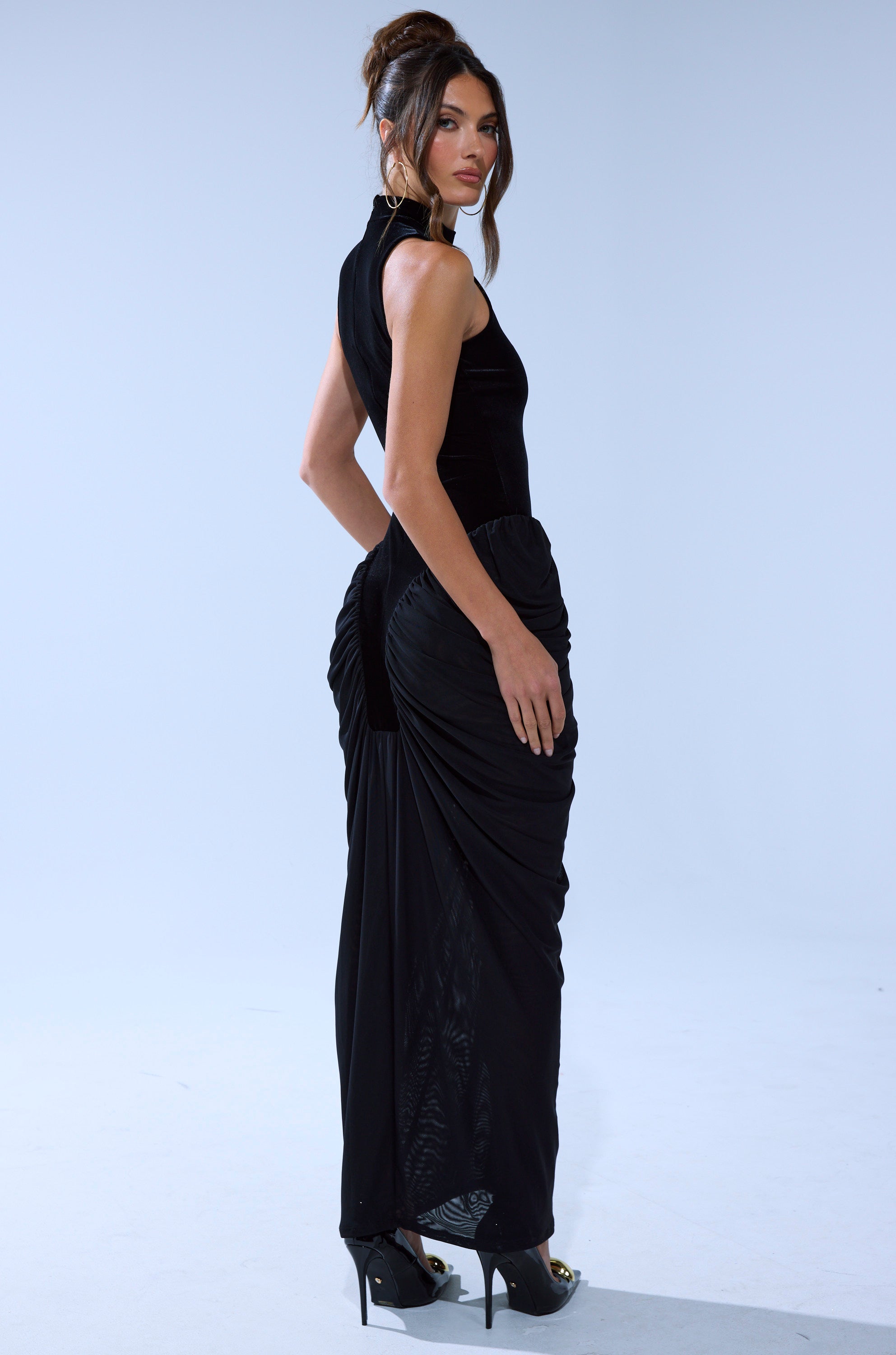 A woman with dark hair in an updo models the OUT FOR THE NIGHT MOCK NECK MAXI DRESS—a black sleeveless, ruched dress—paired with high heels, standing sideways in a minimalist light gray studio.